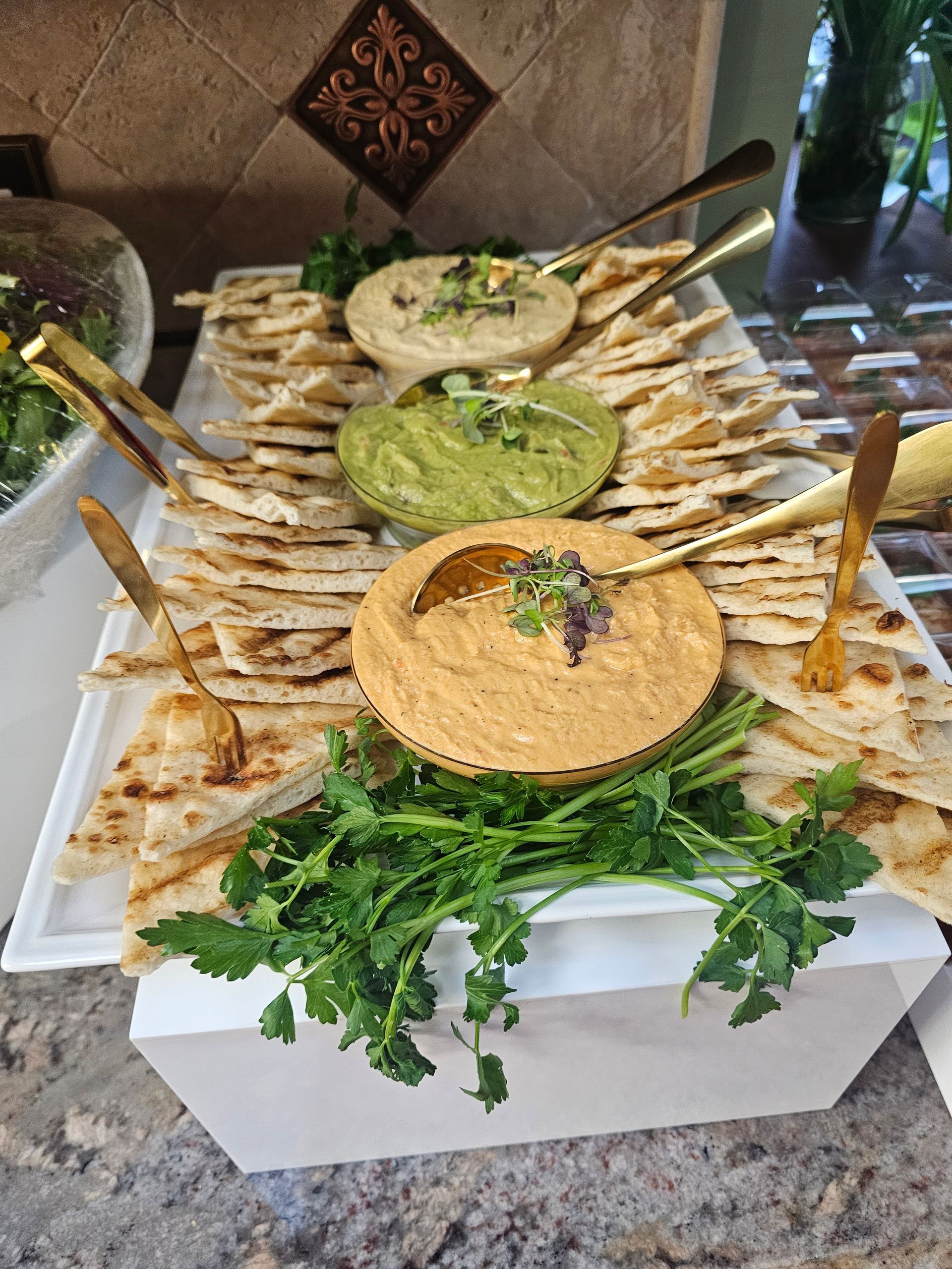 Assorted dips on a platter with pita wedges and herbs on a stone countertop