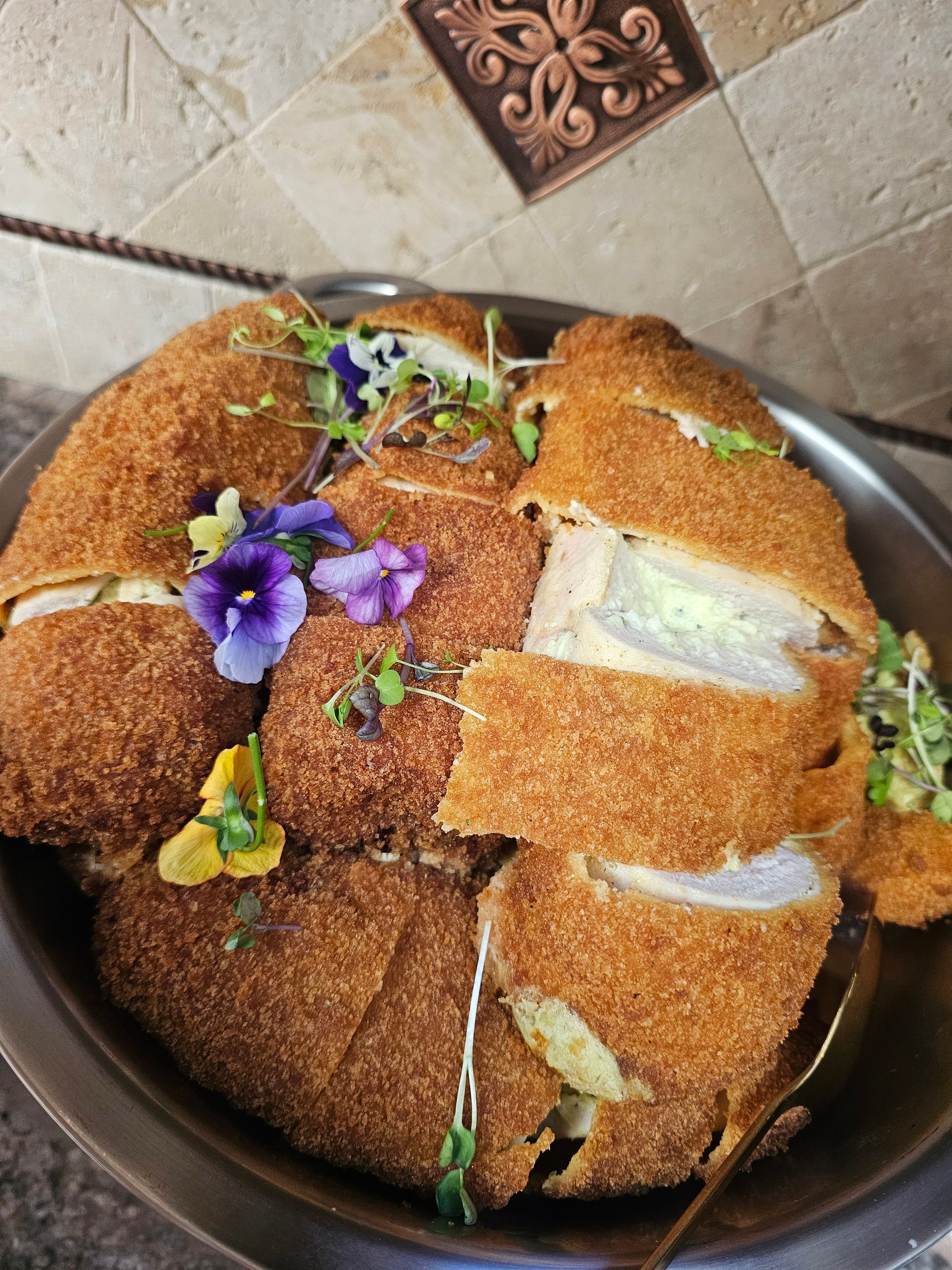 Round loaf of bread topped with purple flowers and green herbs on a dark plate