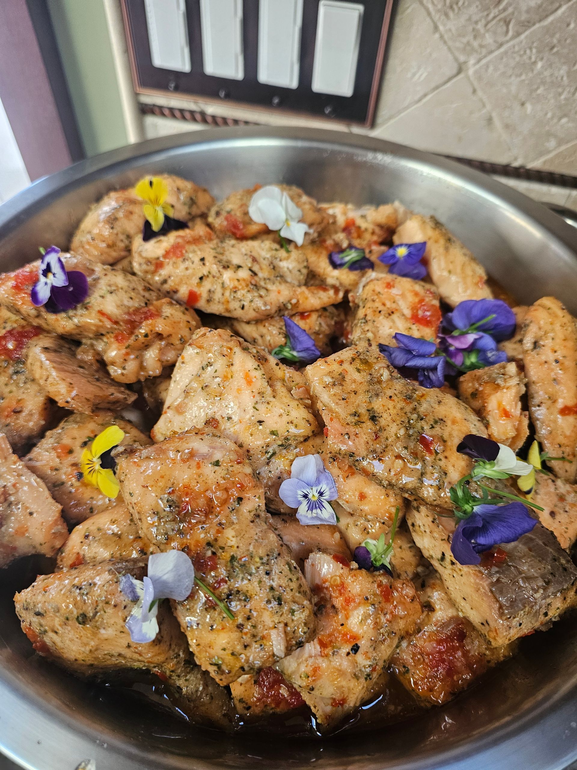Herbed roasted chicken pieces in a metal pan, garnished with purple edible flowers and pepper flakes