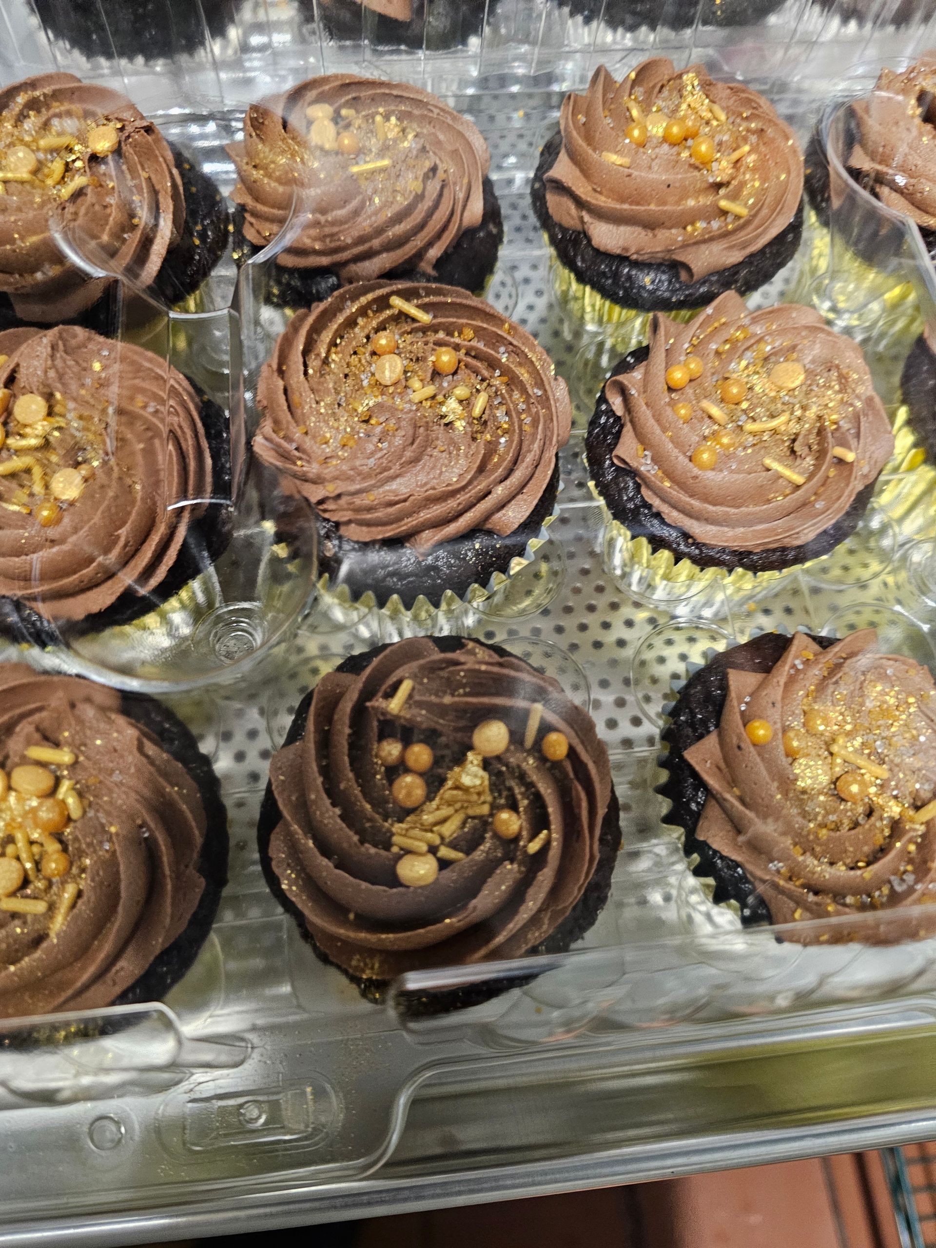 Assorted chocolate cupcakes with swirled frosting and gold sprinkles in a clear plastic tray