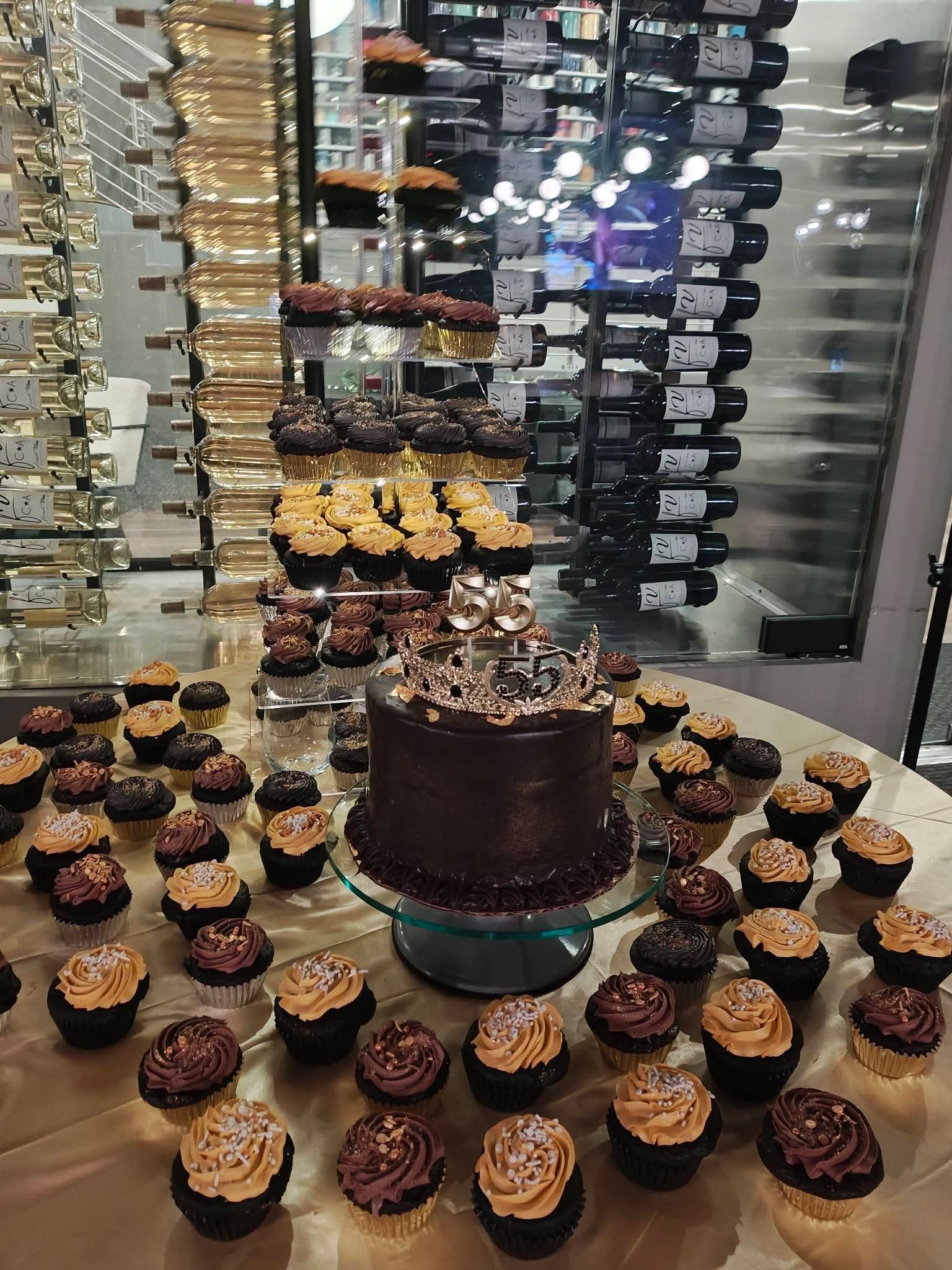 Chocolate cake surrounded by cupcakes on a dessert table in front of a wine display