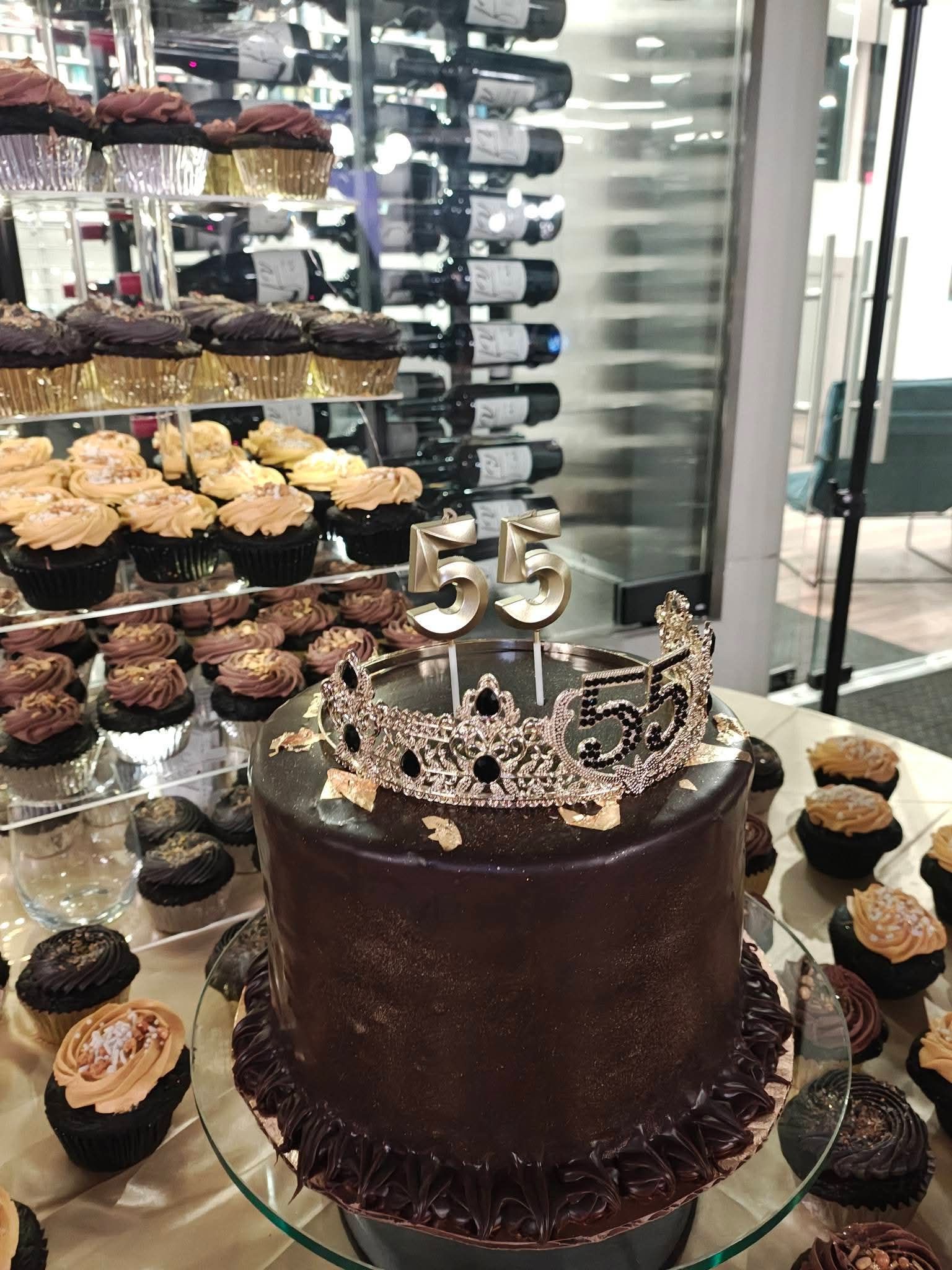 Chocolate cake with crown toppers displayed beside rows of cupcakes in a bakery case