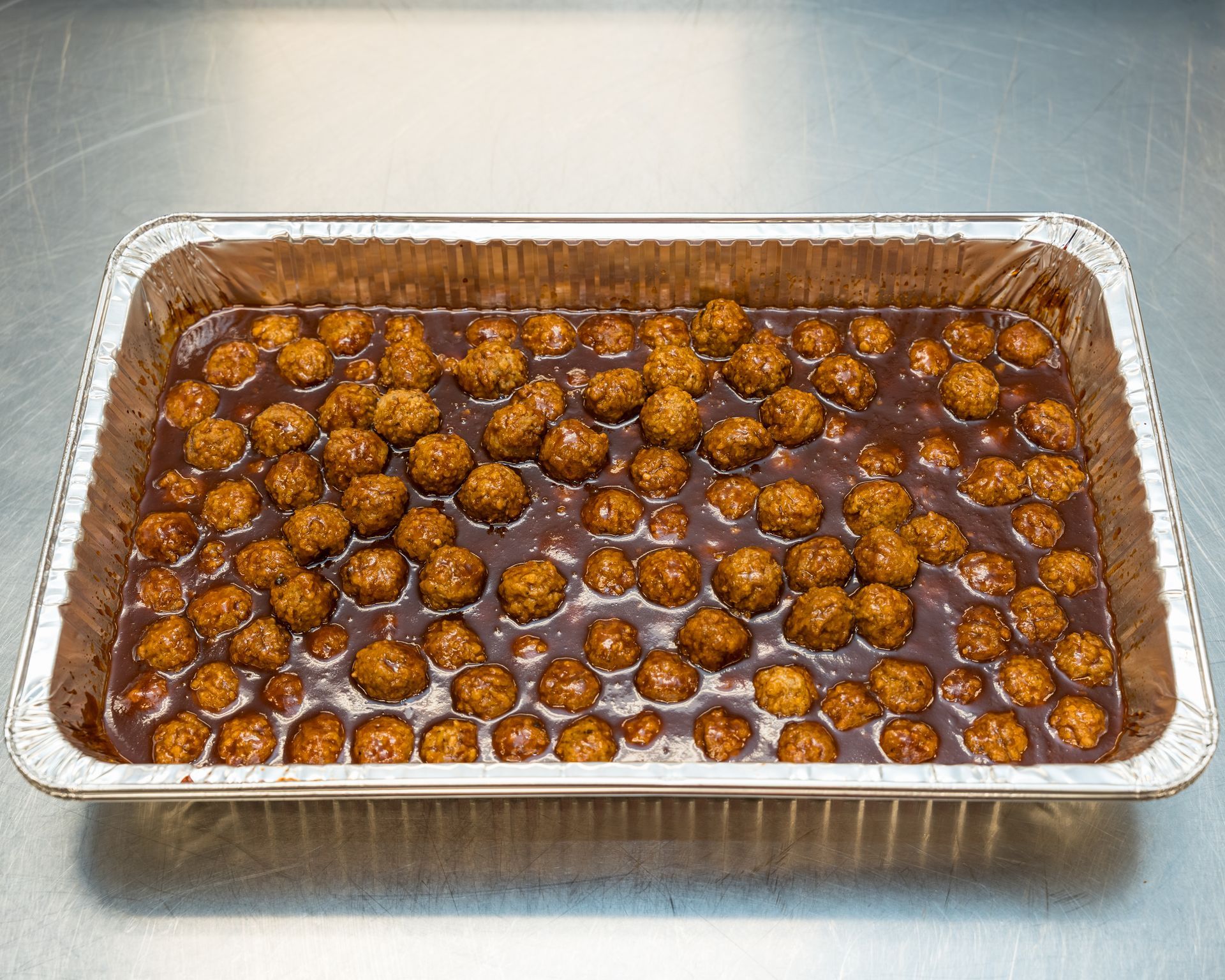 Foil tray of baked meatballs in brown sauce on a stainless steel surface
