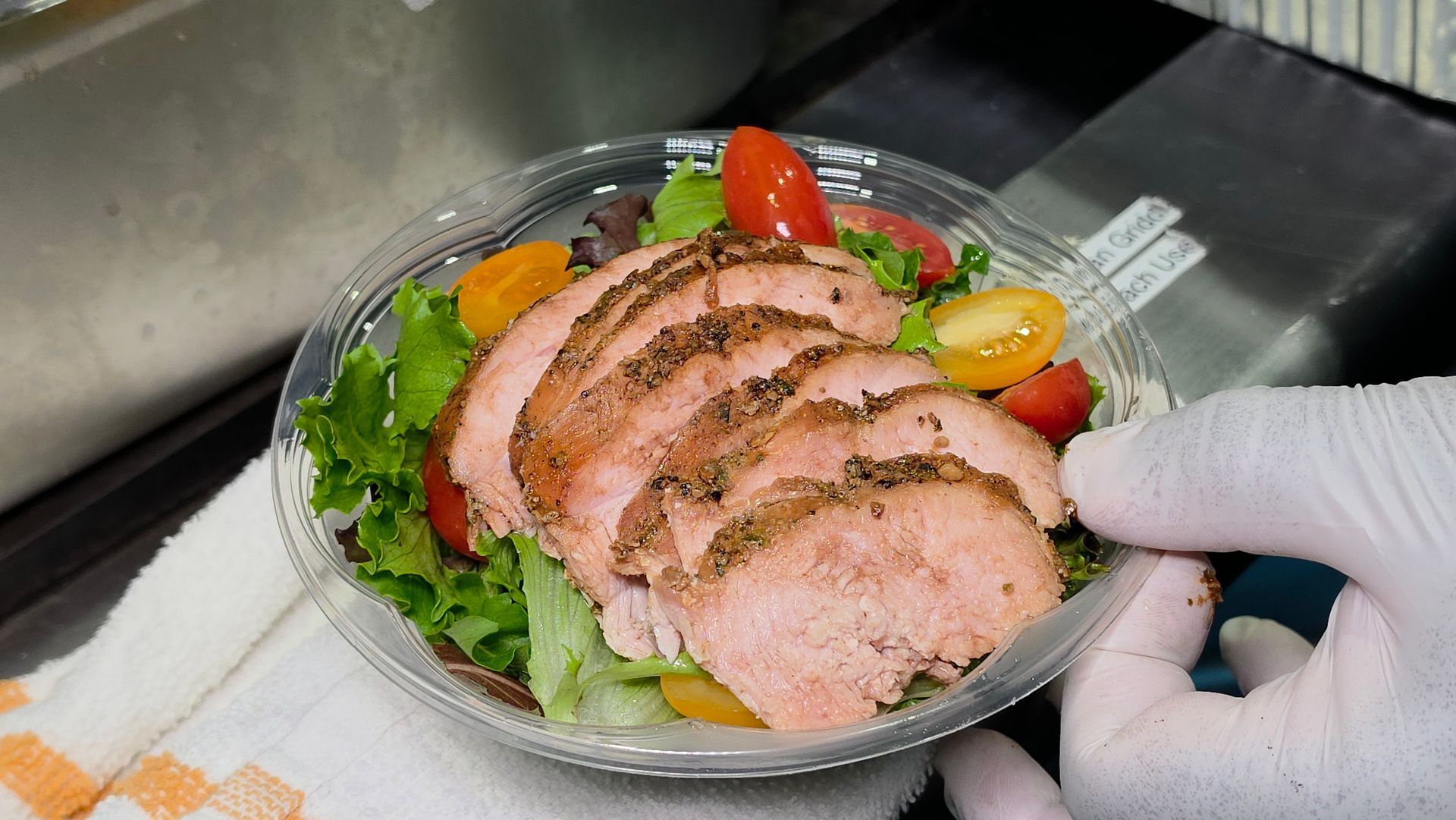 Gloved hand plating sliced seasoned meat over mixed greens and cherry tomatoes in a kitchen bowl