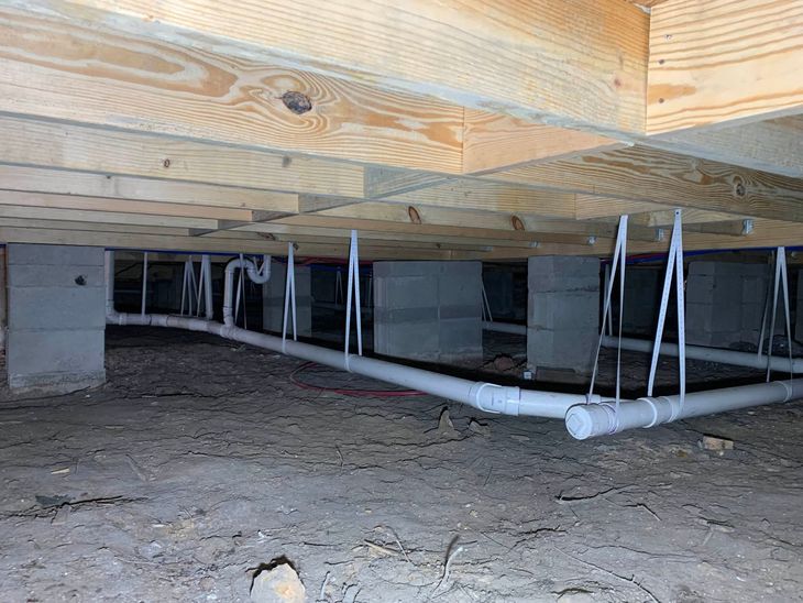Crawlspace under a wooden floor with exposed white pipes and supports over dirt ground