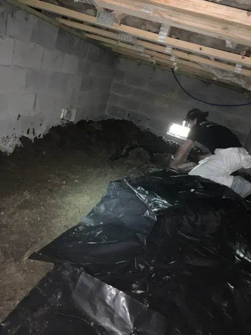Person working in a dim crawlspace with black plastic sheeting and exposed pipes on the ceiling