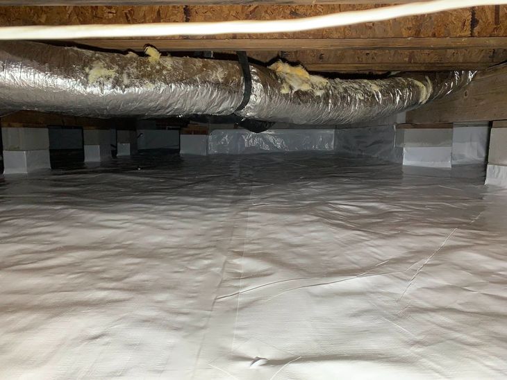 Crawlspace with silver foil insulation wrapped around ducts above a white vapor barrier floor