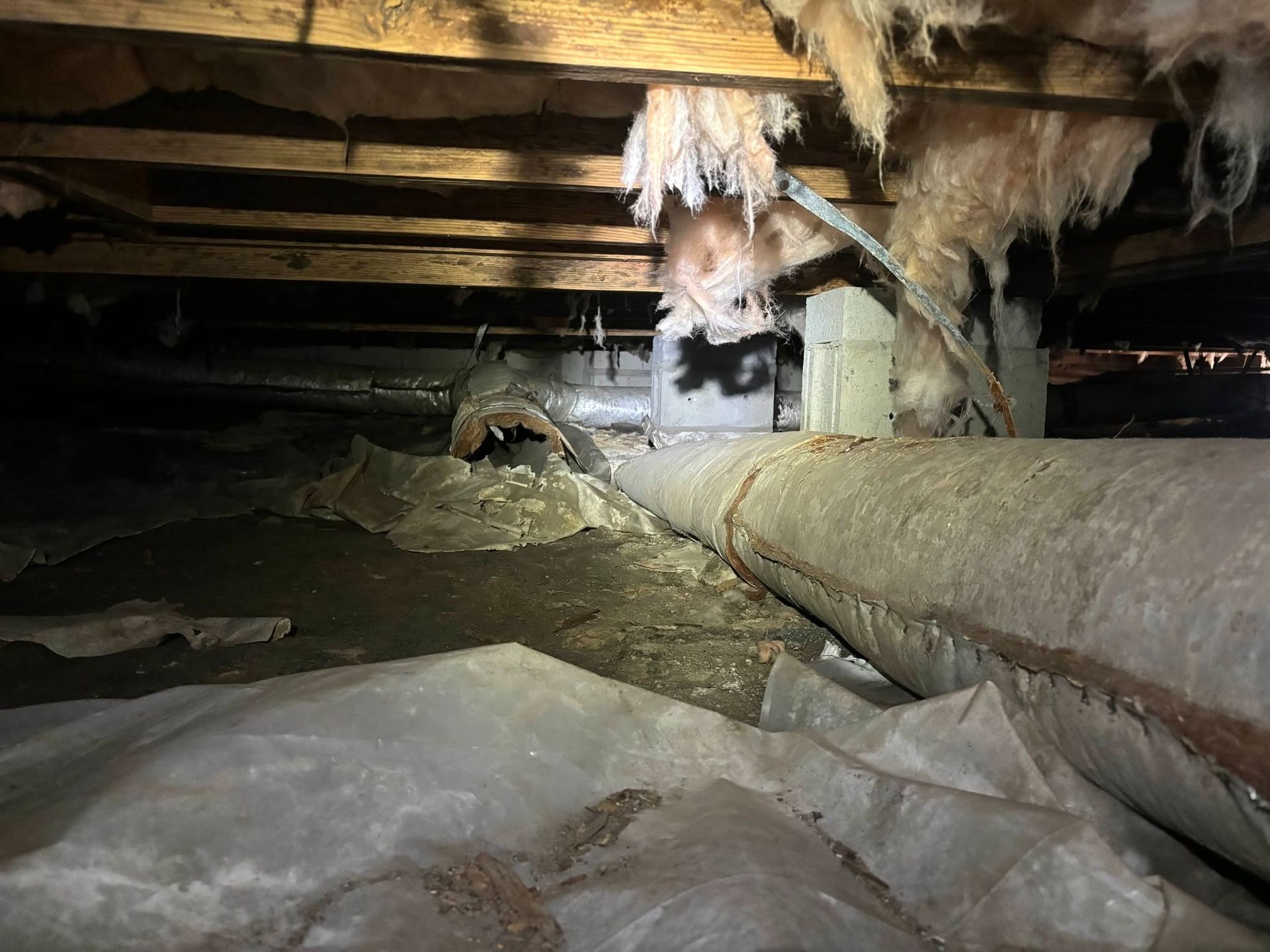 Crawlspace with exposed pipes, beams, and insulation, dimly lit with damp, muddy ground.