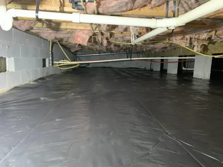 Crawlspace with black vapor barrier flooring, exposed insulation, ducts, and support beams under a house
