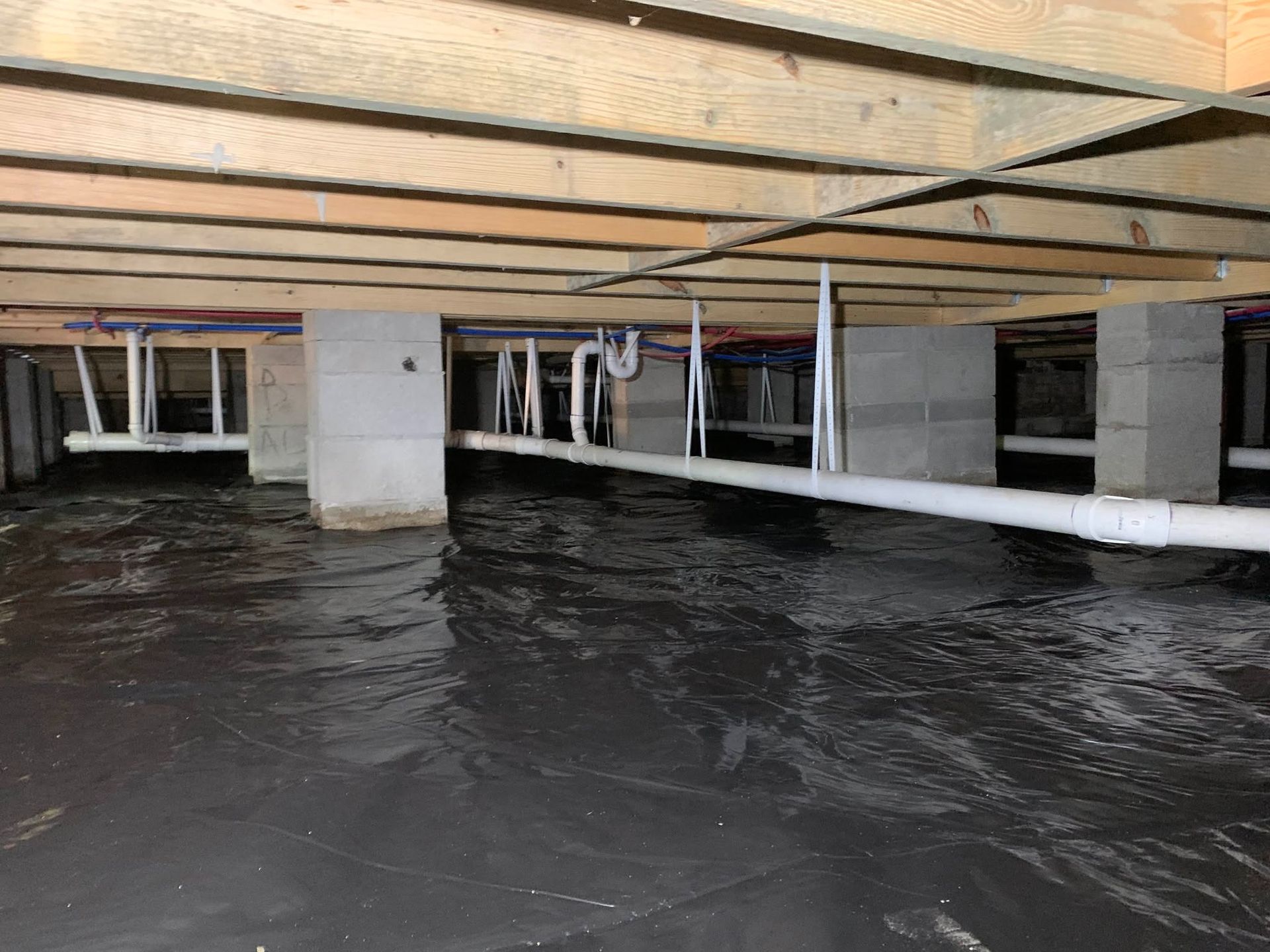 Flooded crawl space under a house with exposed beams, pipes, and standing water
