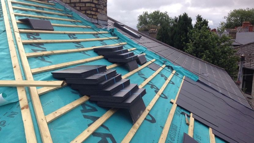Roof under construction with blue underlayment, wooden beams, and stacked dark tiles.
