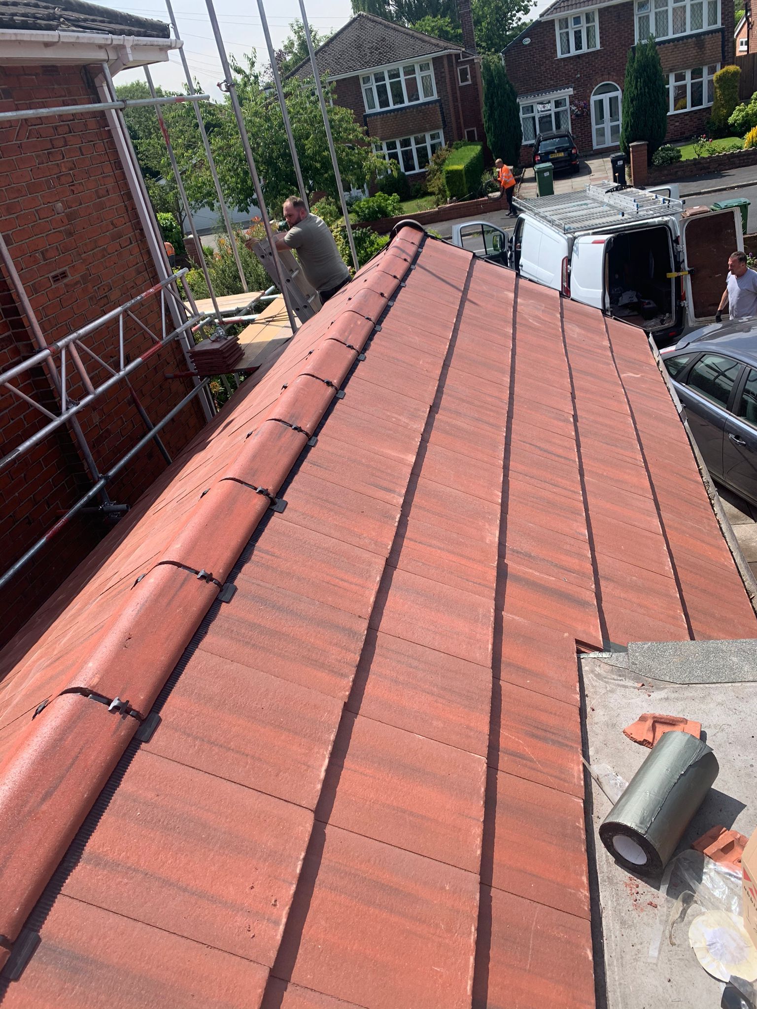 Red tile roof with a person standing on it, construction site with scaffolding and houses in the background.