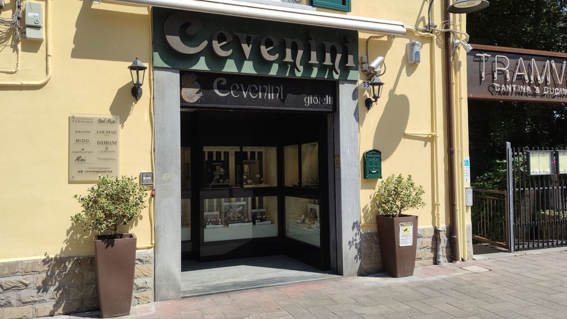 Ingresso del negozio con tenda da sole nera, piante in vaso e un vialetto piastrellato di fronte a un edificio beige.