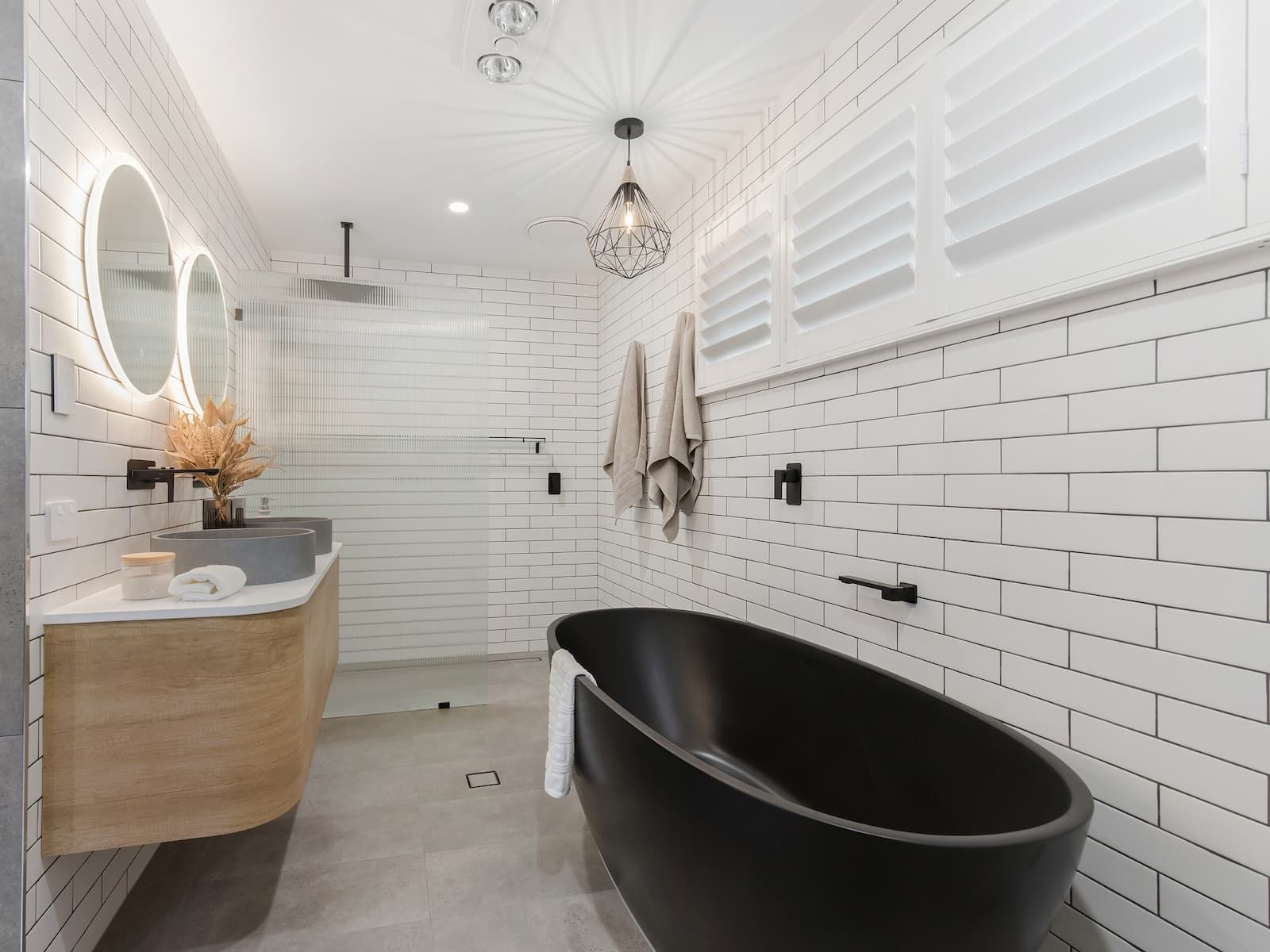 Modern bathroom with black soaking tub, wood vanity, white brick-style tile, and round mirrors.