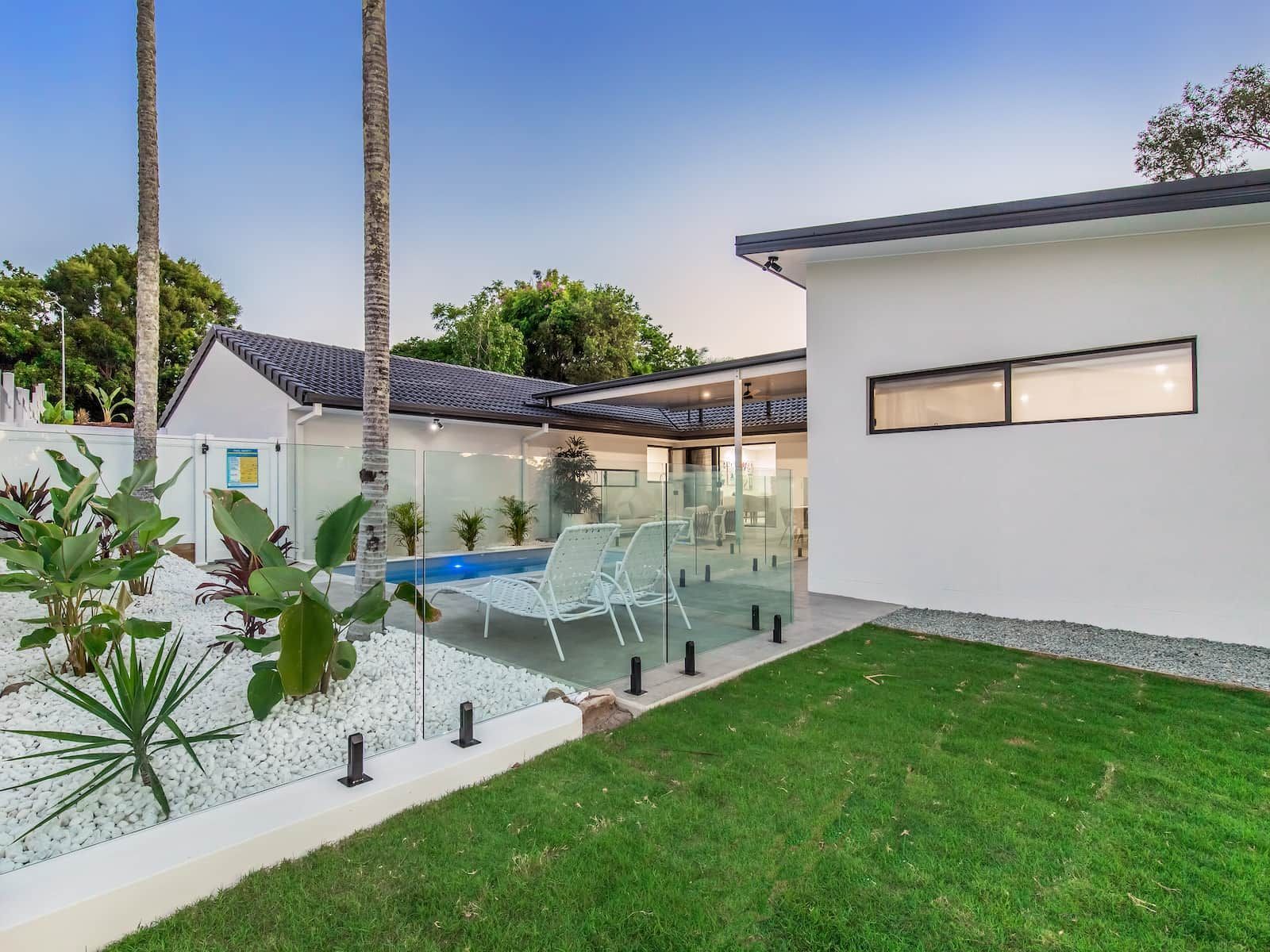White house exterior with pool, glass fence, palm trees, and green lawn.