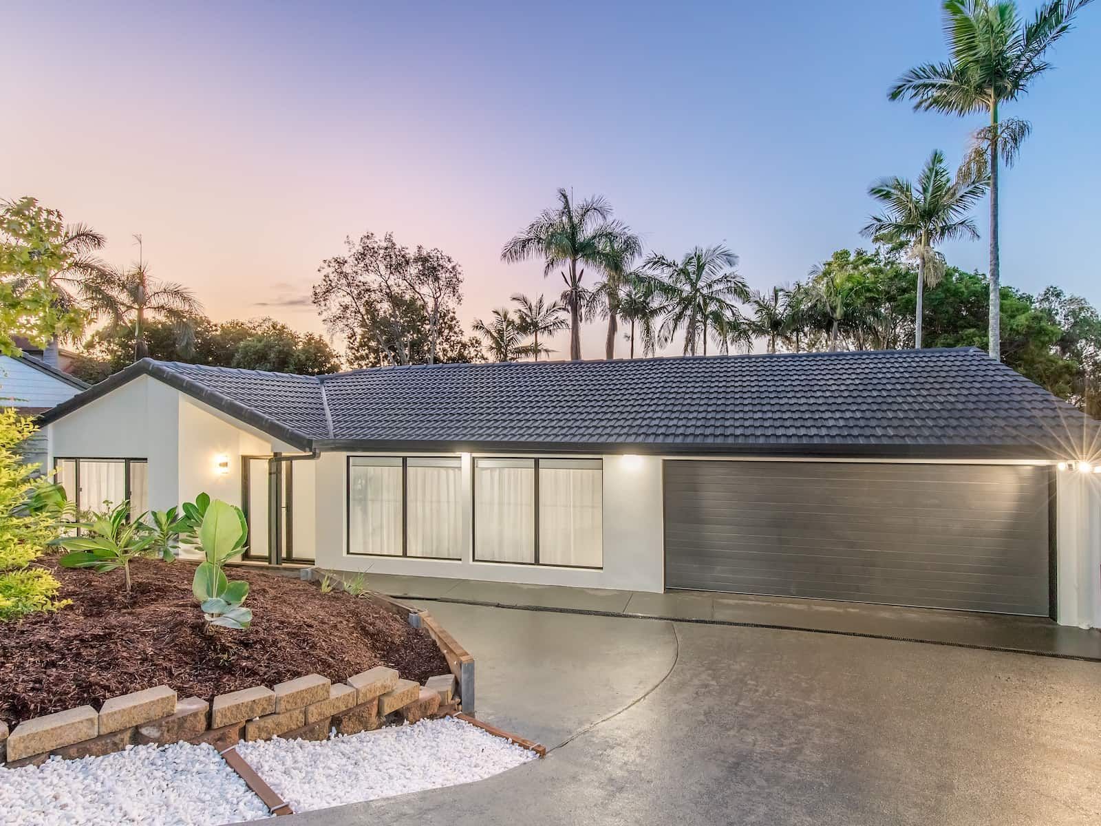 Modern, single-story house with gray roof and garage, palm trees, and driveway.