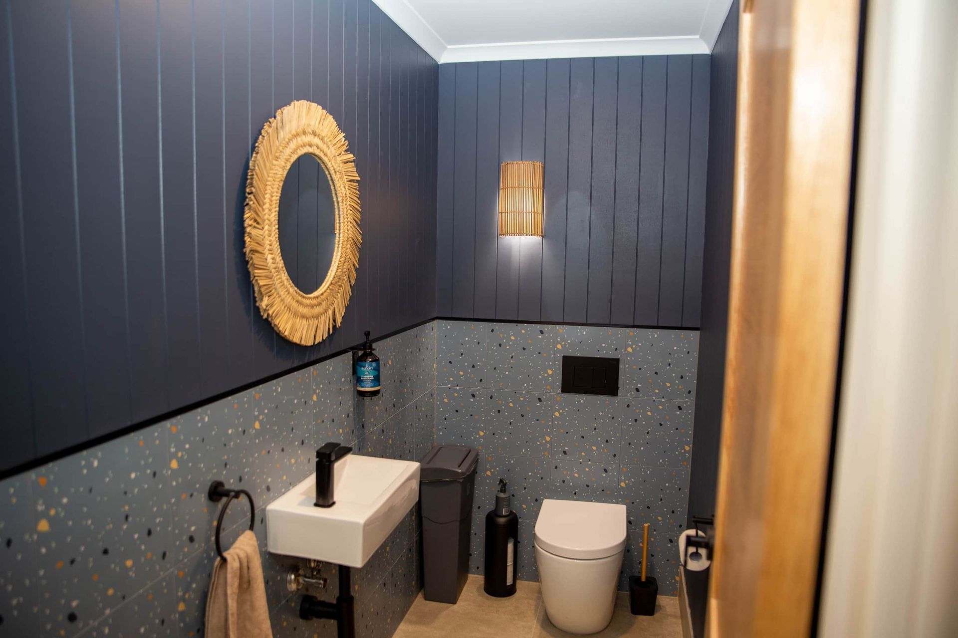 A modern bathroom with blue paneled walls, a gold mirror, and a terrazzo-tiled lower wall.