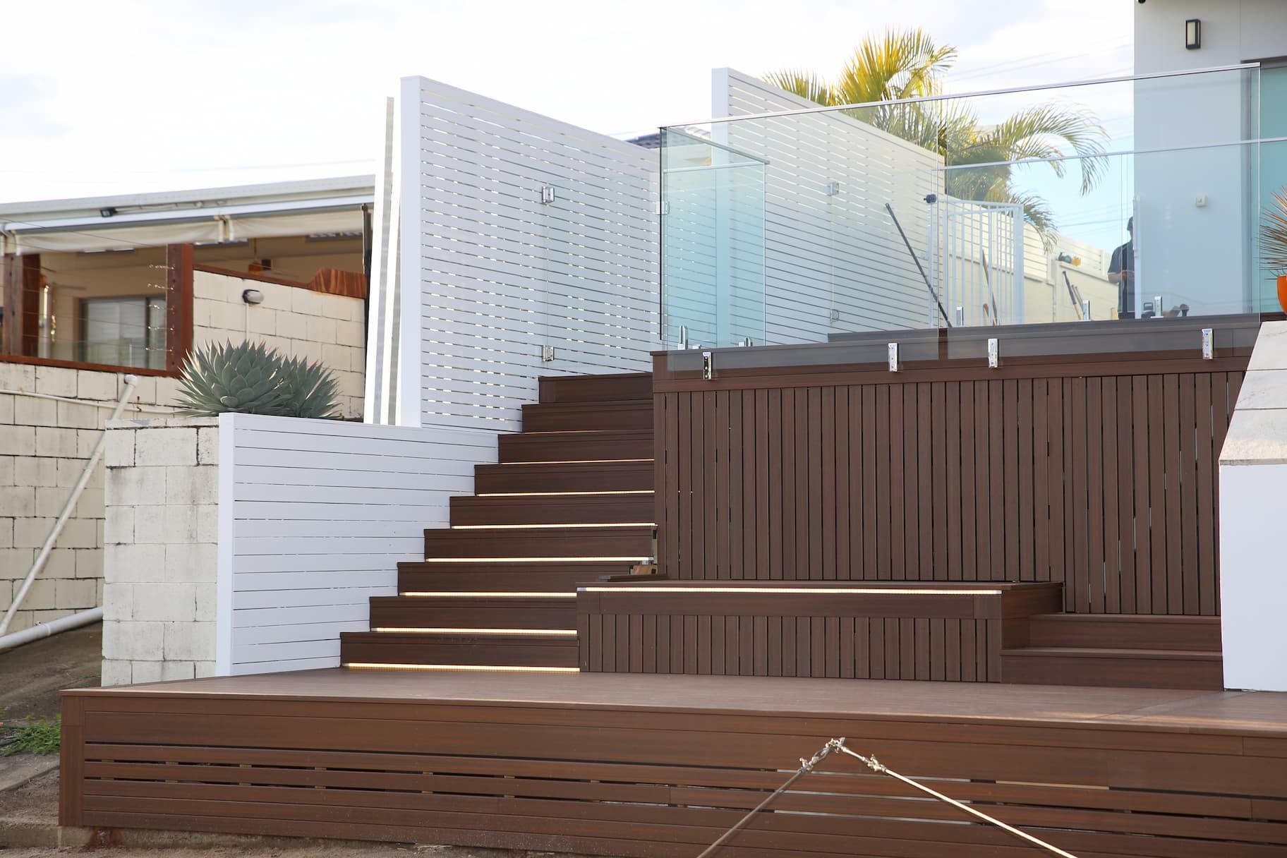 Wooden deck with stairs and white patterned privacy screen, glass railing, and a view of a house.