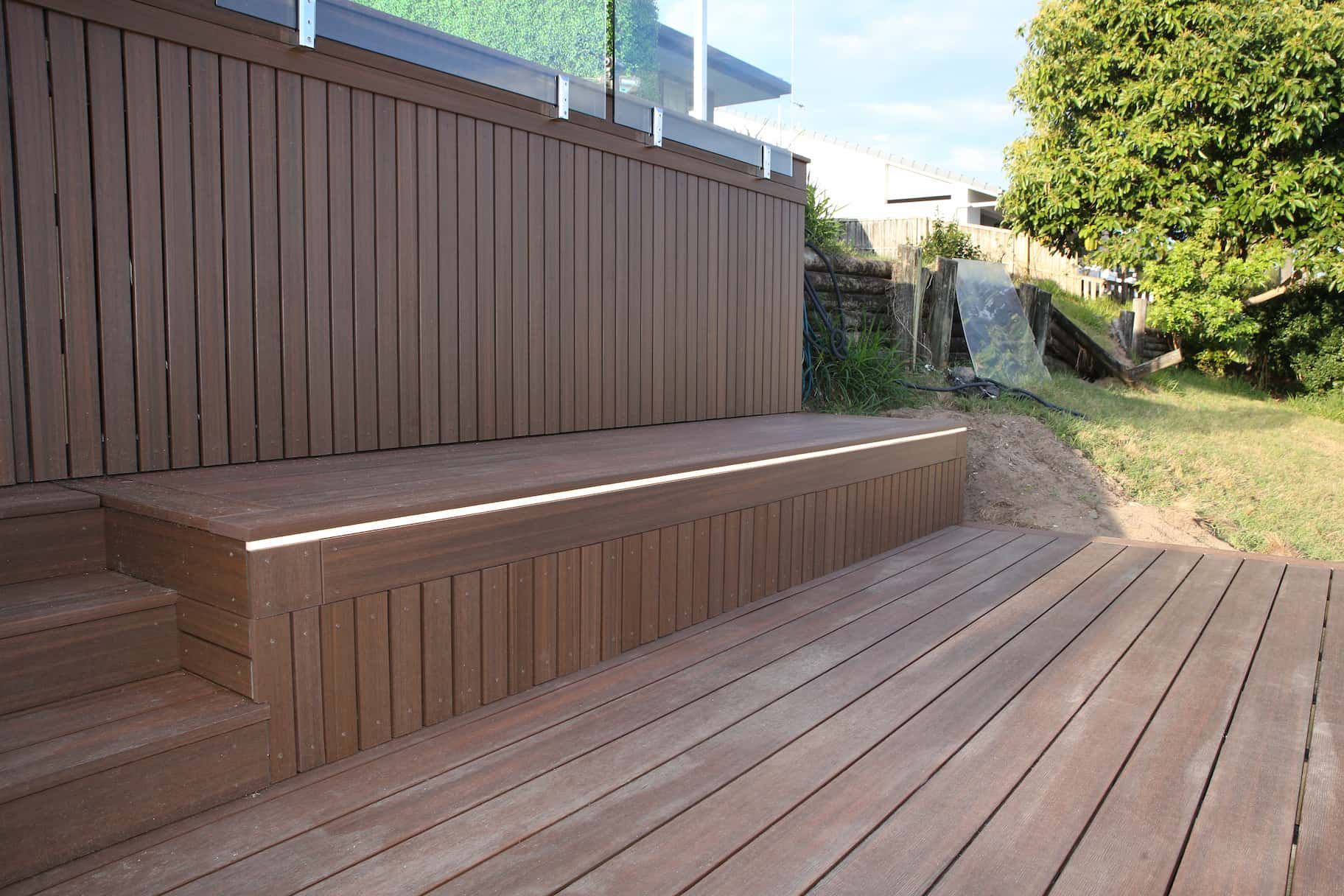 Brown composite deck with steps and vertical paneling against a green lawn and vegetation.