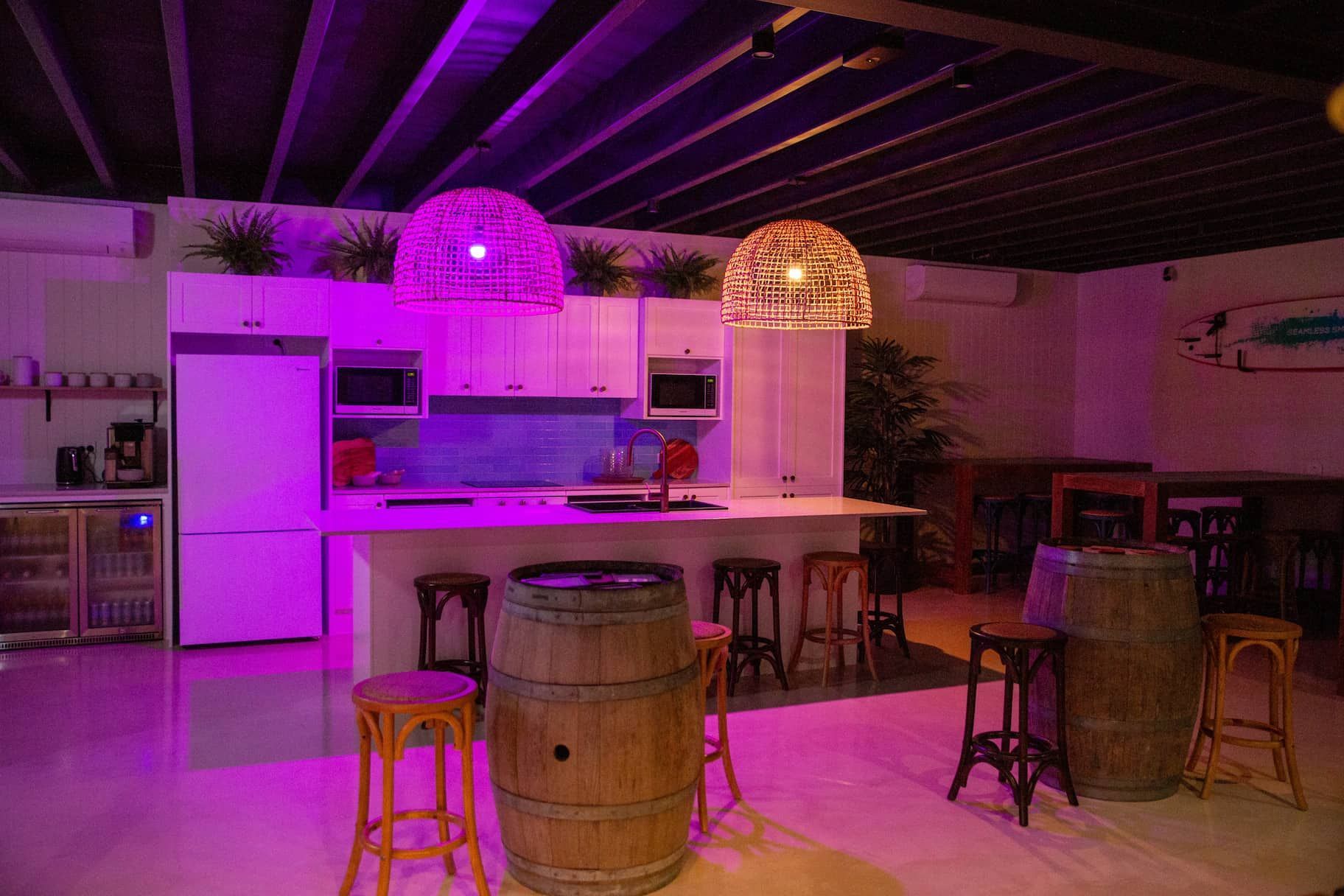 A purple-lit bar and kitchen area with a counter, stools, and barrel tables.