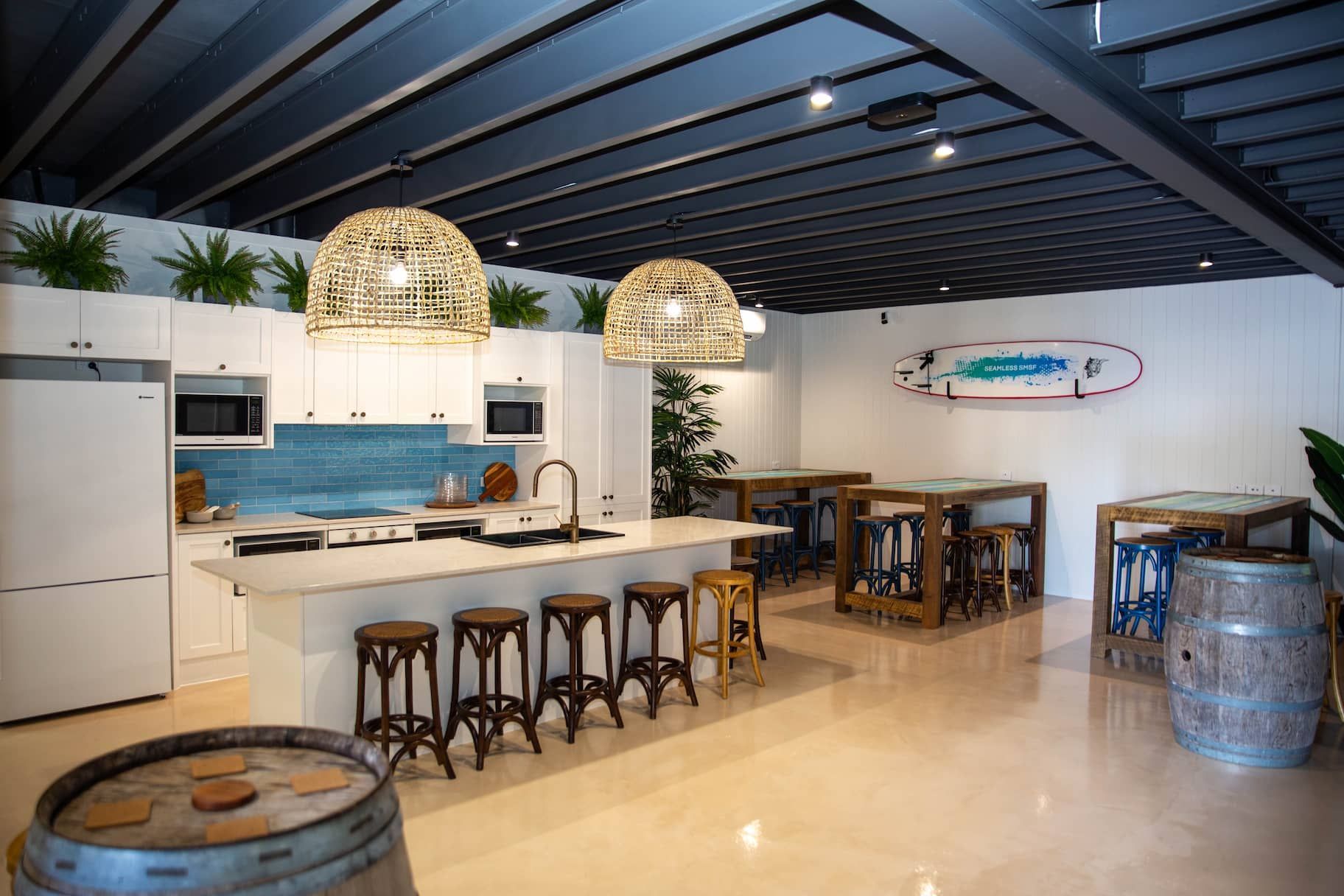 Spacious, modern kitchen with white cabinets, large island, and bar stools. Includes surfboard decor and wood barrel tables.