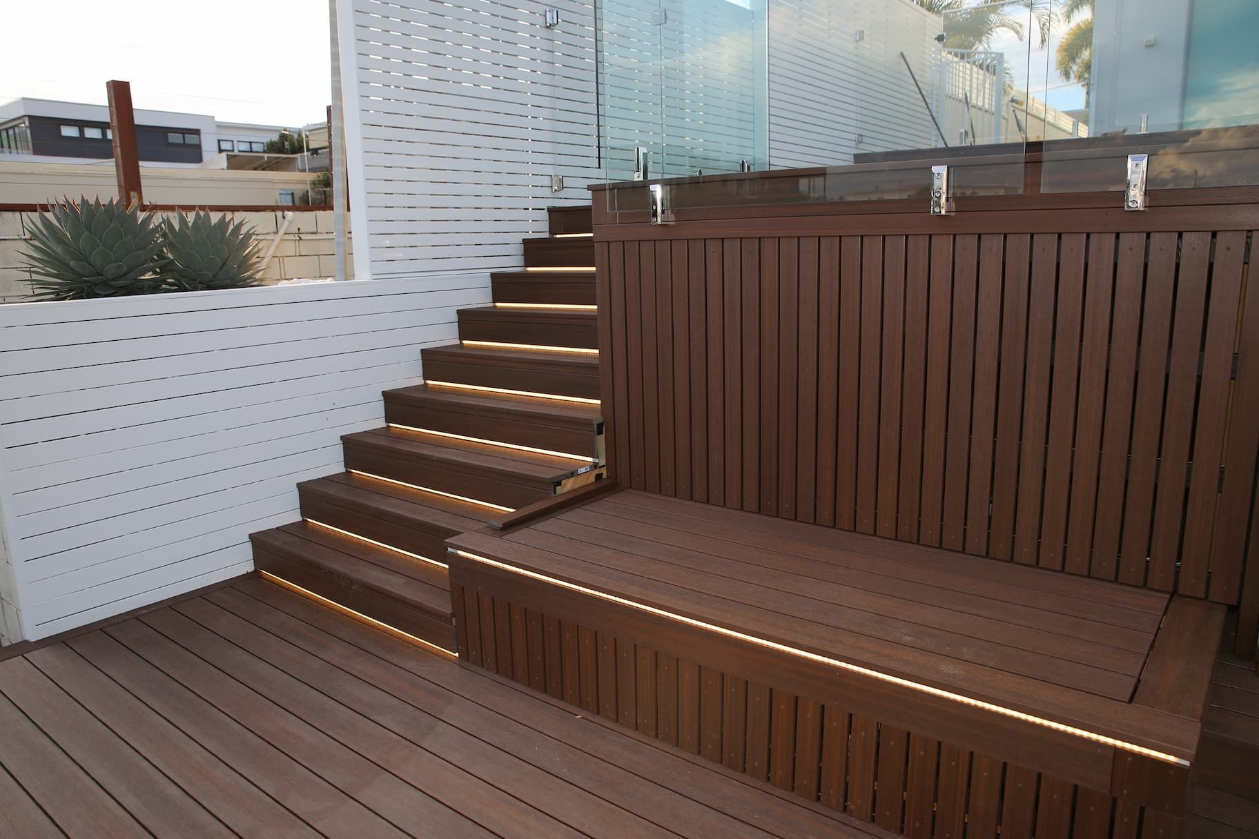 Wooden deck with steps, built-in bench, and accent lighting. White slatted fence and glass wall in background.