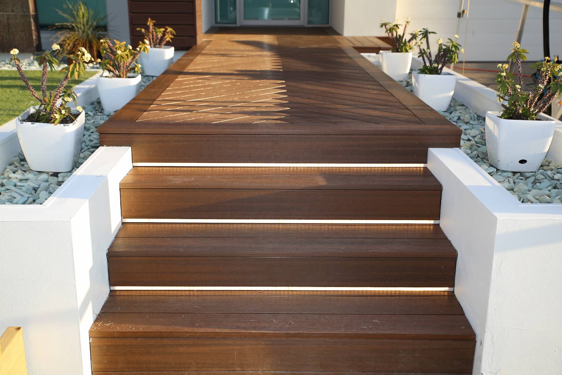 Wooden steps and walkway leading to a house entrance, bordered by white planters with greenery and light-colored rocks.