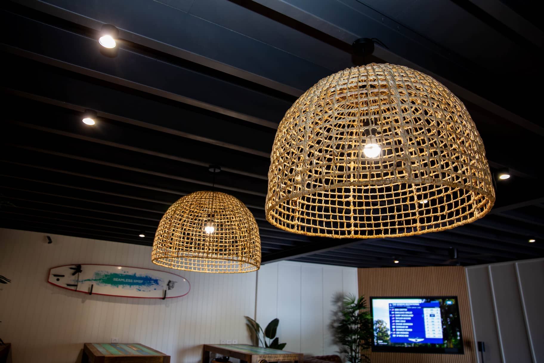 Two woven pendant lights hang above a room with a surfboard and TV on the wall.