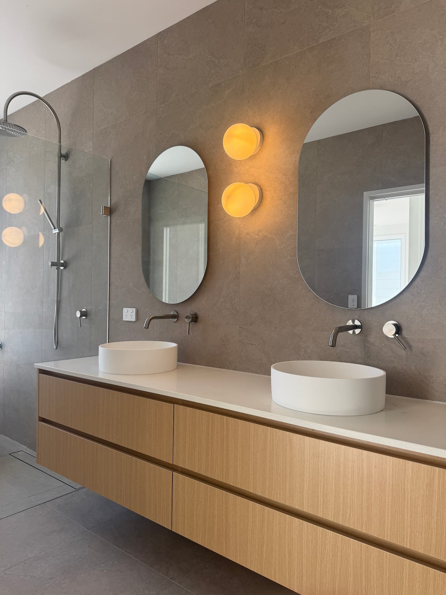 Modern bathroom with light wood vanity, oval mirrors, two vessel sinks, and gray tile walls.