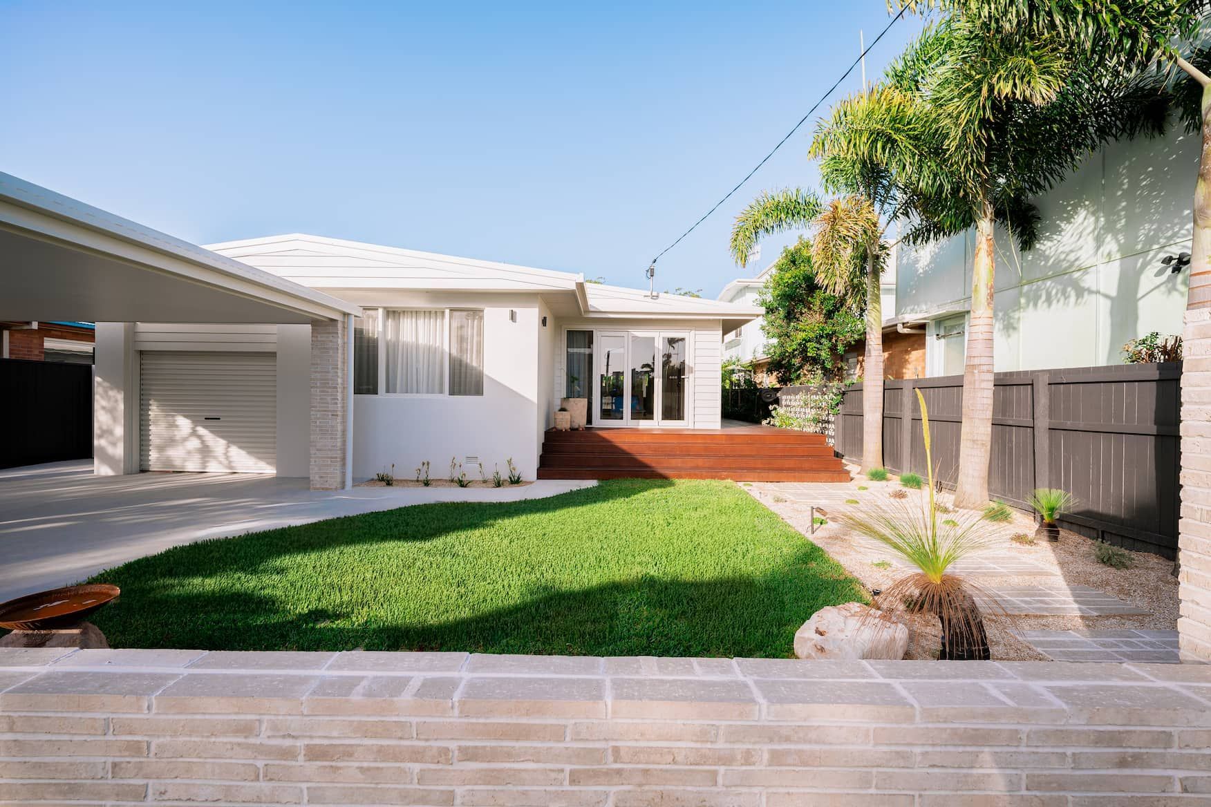 White house with carport, wooden deck, green lawn, palm trees, and gray fence.
