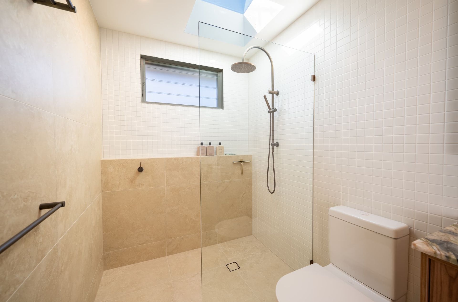 Bathroom with white tile walls, glass shower door, and toilet. Skylight and small window present.