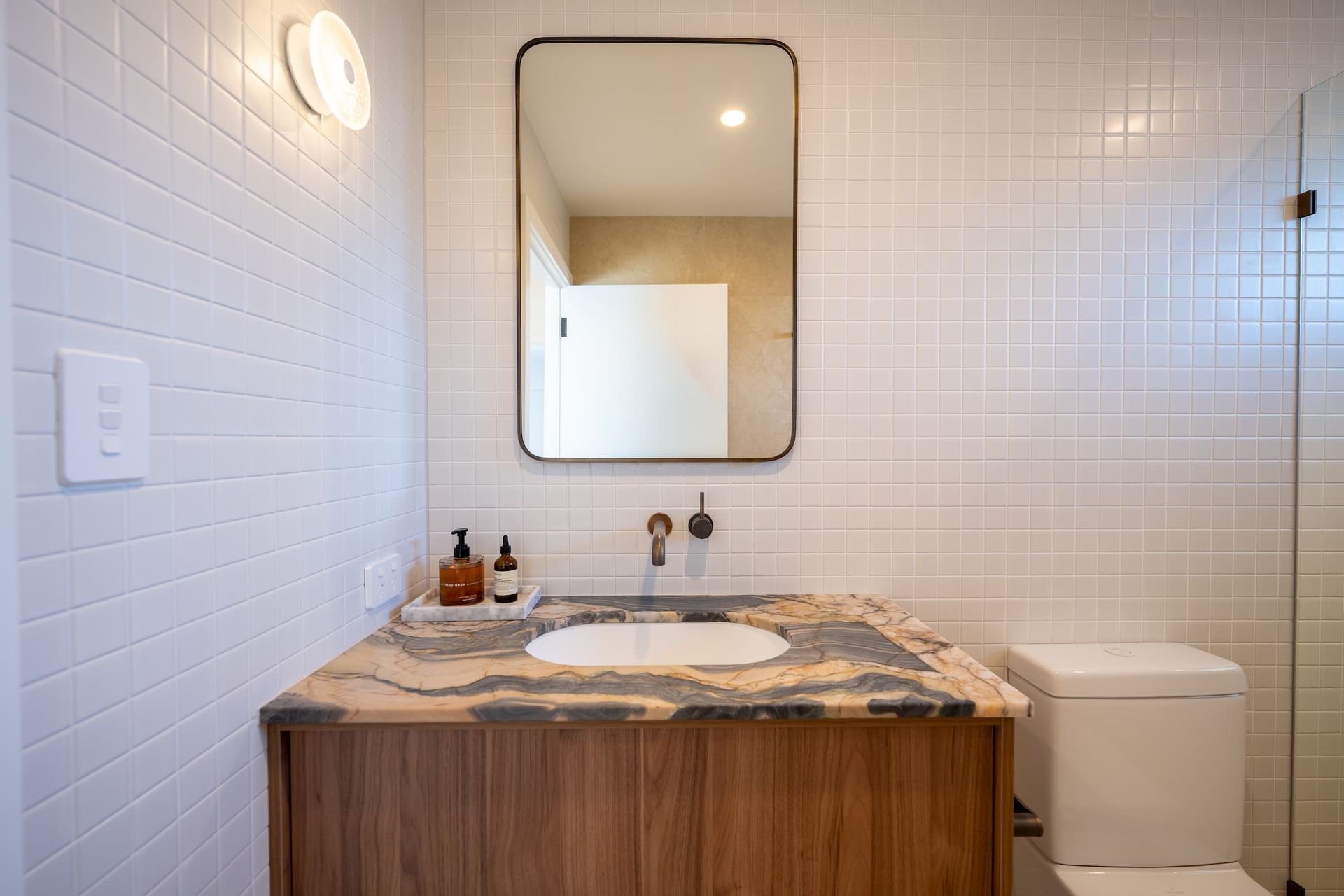 Bathroom with wooden vanity, marble countertop, rectangular mirror, and white tiled walls.
