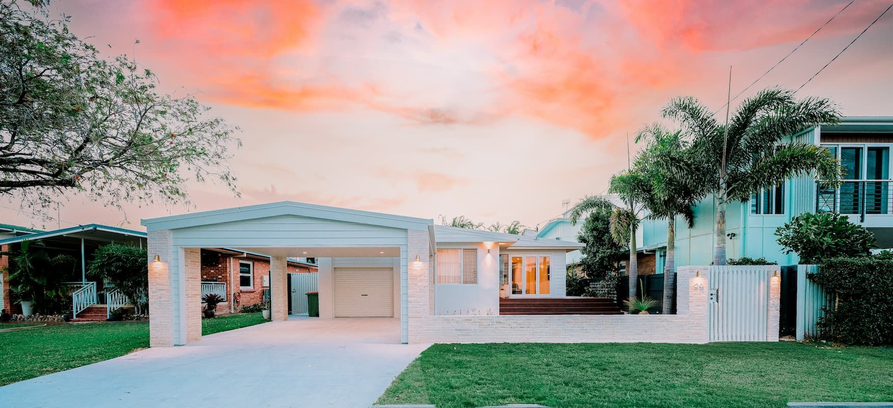 White house with a carport and garage, green lawn, palm trees and orange sky.