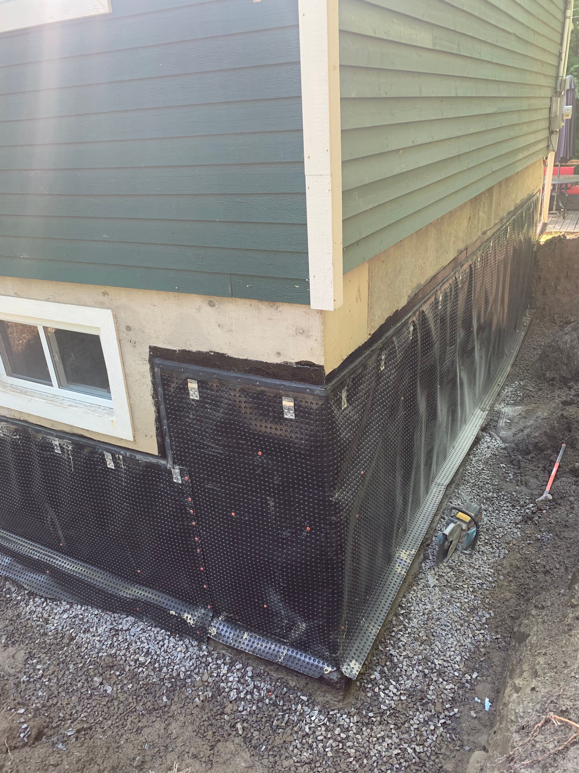 Un mur de fondation avec une membrane de drainage noire à picots installée, partiellement recouverte de gravier dans une tranchée.