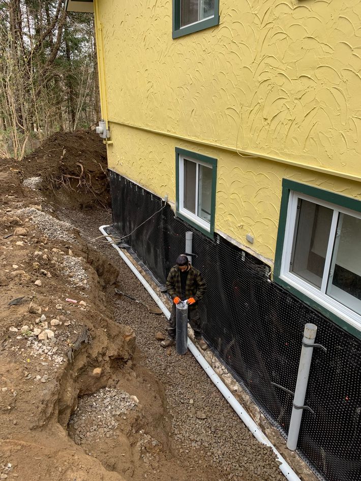 Un ouvrier installe un tuyau de drainage le long des fondations étanches en noir d'une maison jaune lors de travaux de rénovation extérieure.