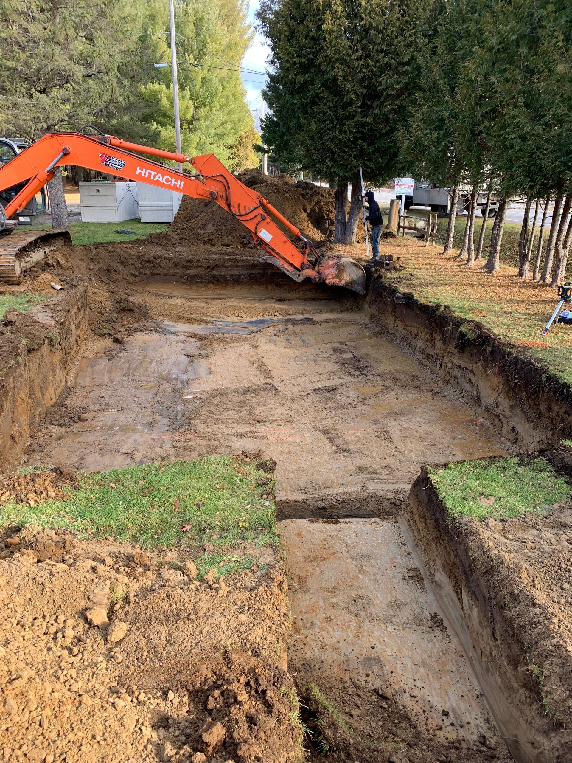 Une pelle mécanique Hitachi orange travaille dans une tranchée boueuse, profonde et rectangulaire, sur un terrain herbeux près des arbres.
