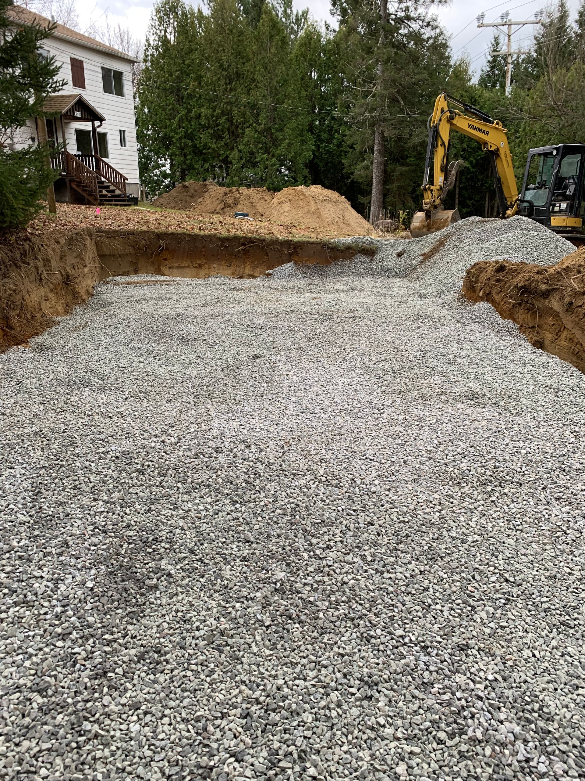 Un chantier de terrassement rempli de gravier, avec une excavatrice jaune en arrière-plan, près d'une maison.
