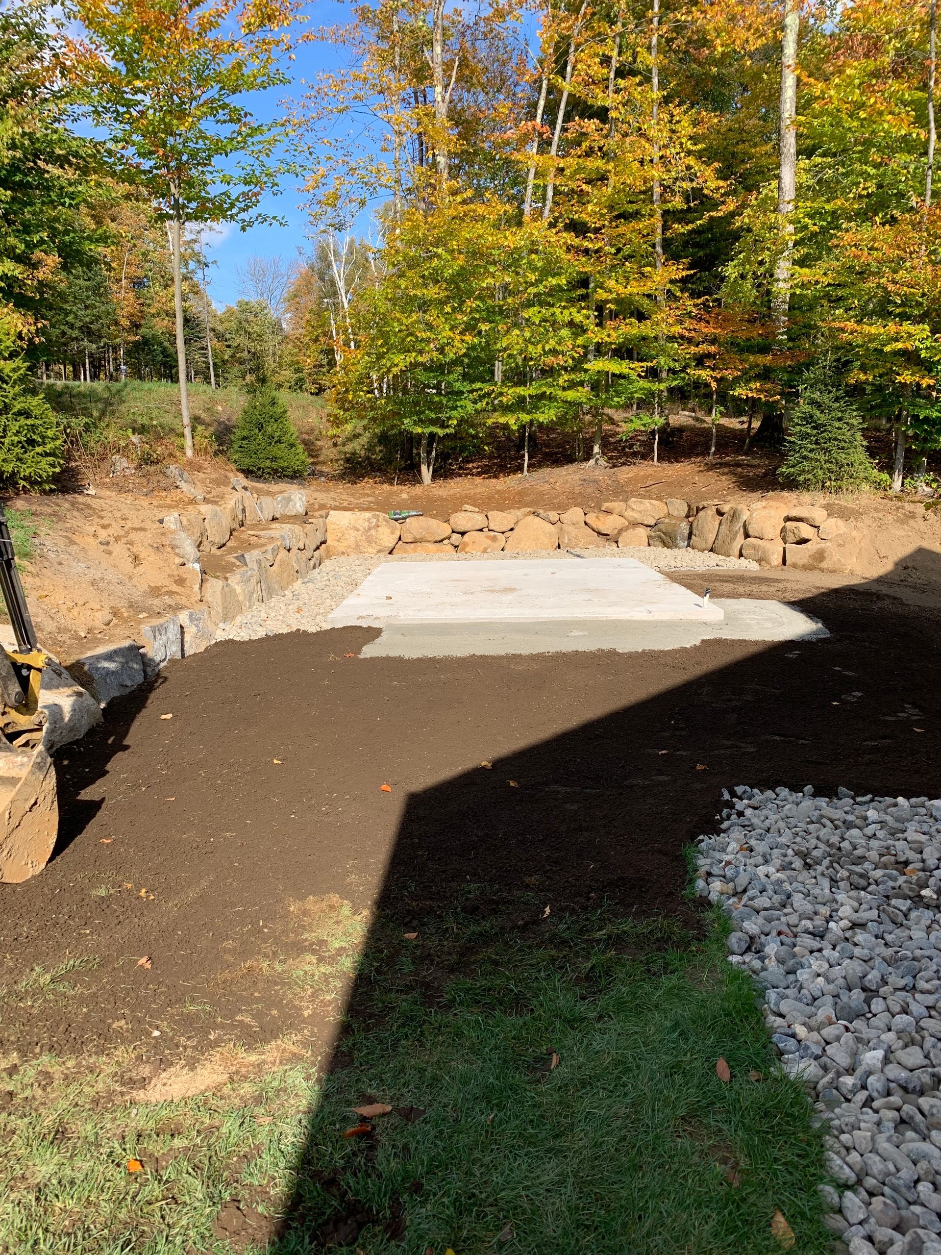 Une dalle de béton gris clair, enfouie dans un sol sombre et frais recouvert de gravier, bordée par un mur de soutènement en pierre et des arbres aux couleurs d'automne.