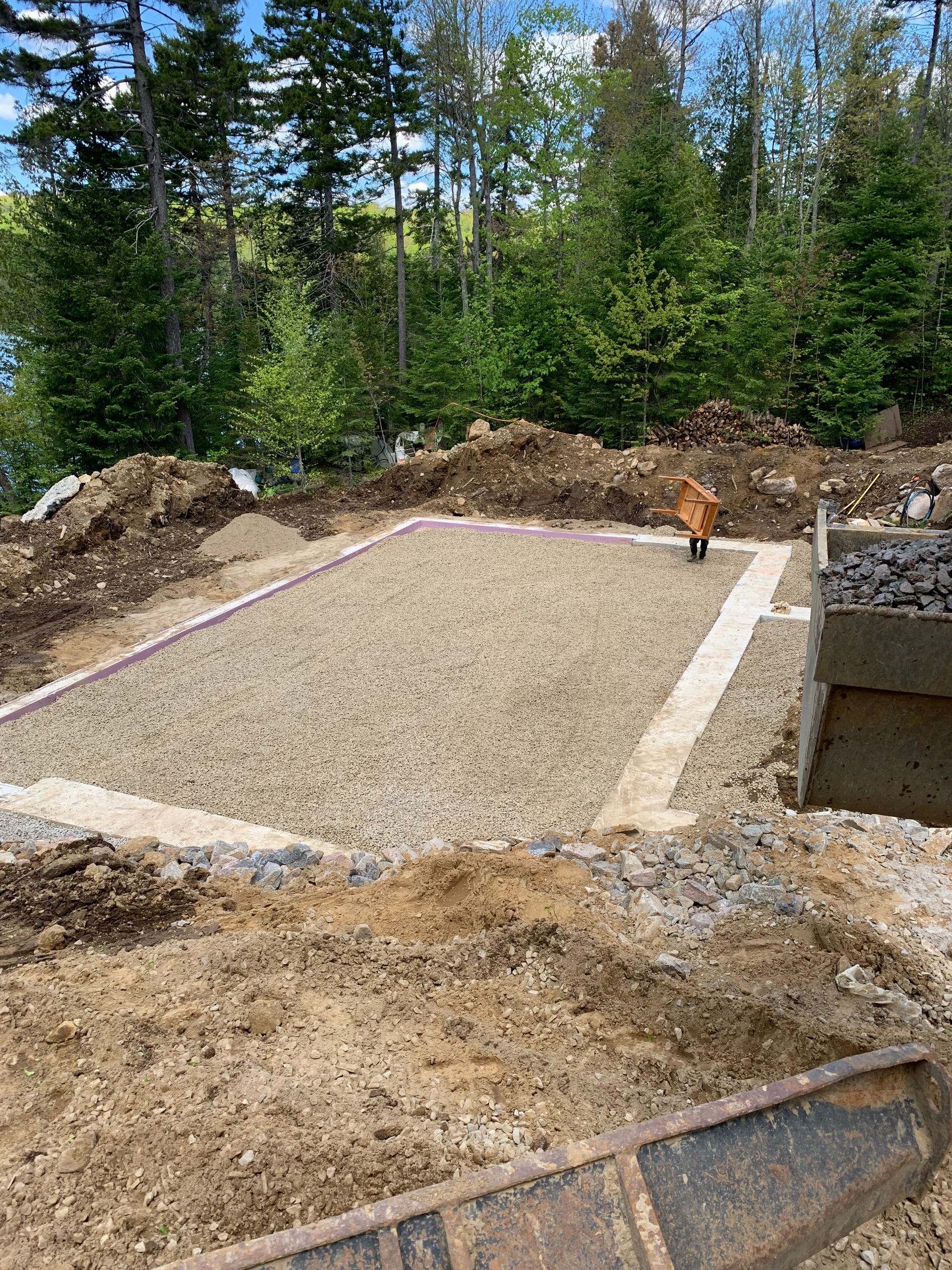 Une fondation rectangulaire en gravier, destinée à accueillir une structure, se trouve dans une clairière entourée d'une forêt.