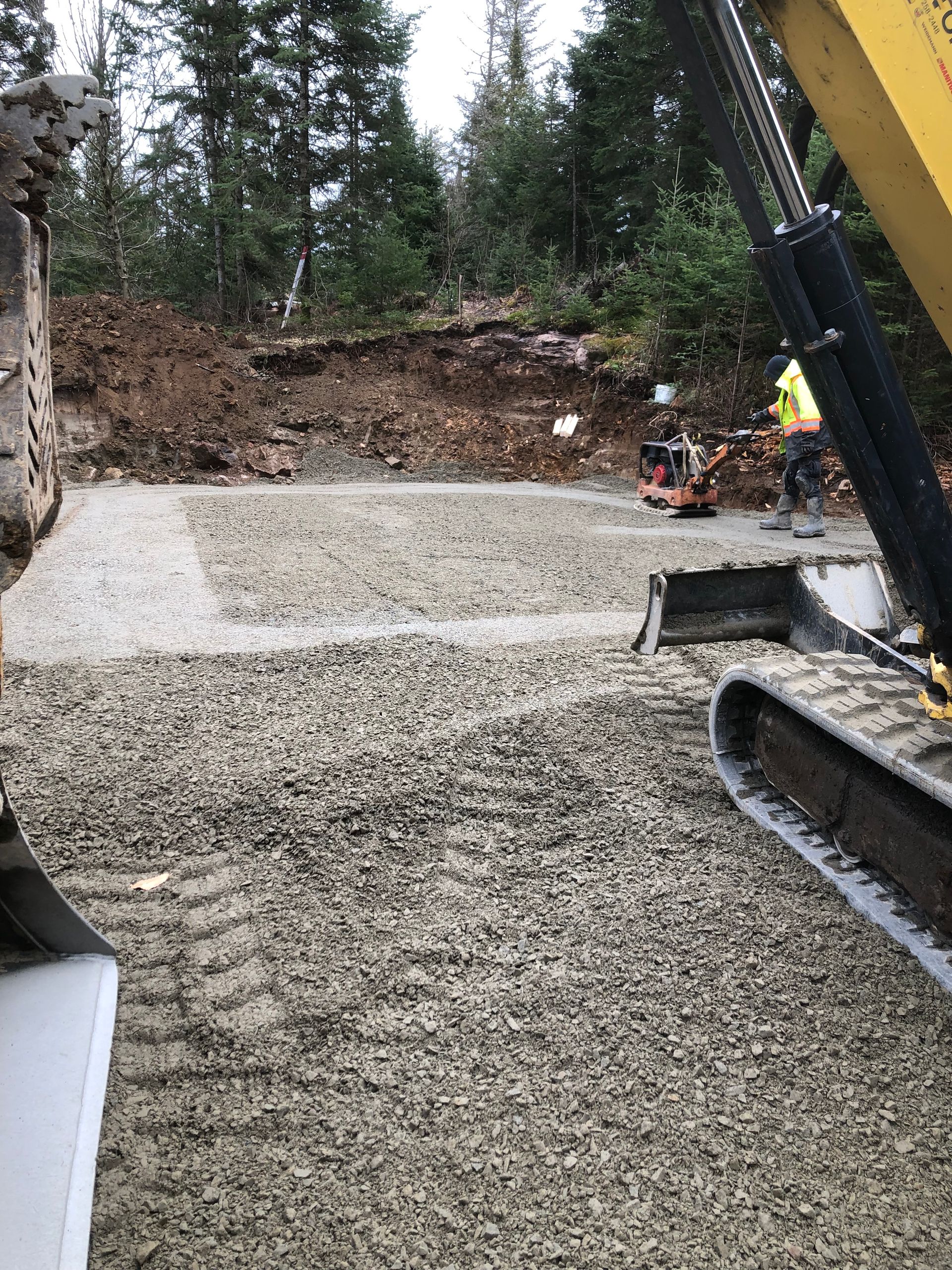 Une excavatrice est garée à côté d'un chantier de construction en gravier fraîchement nivelé, dégagé au pied d'une colline boisée.