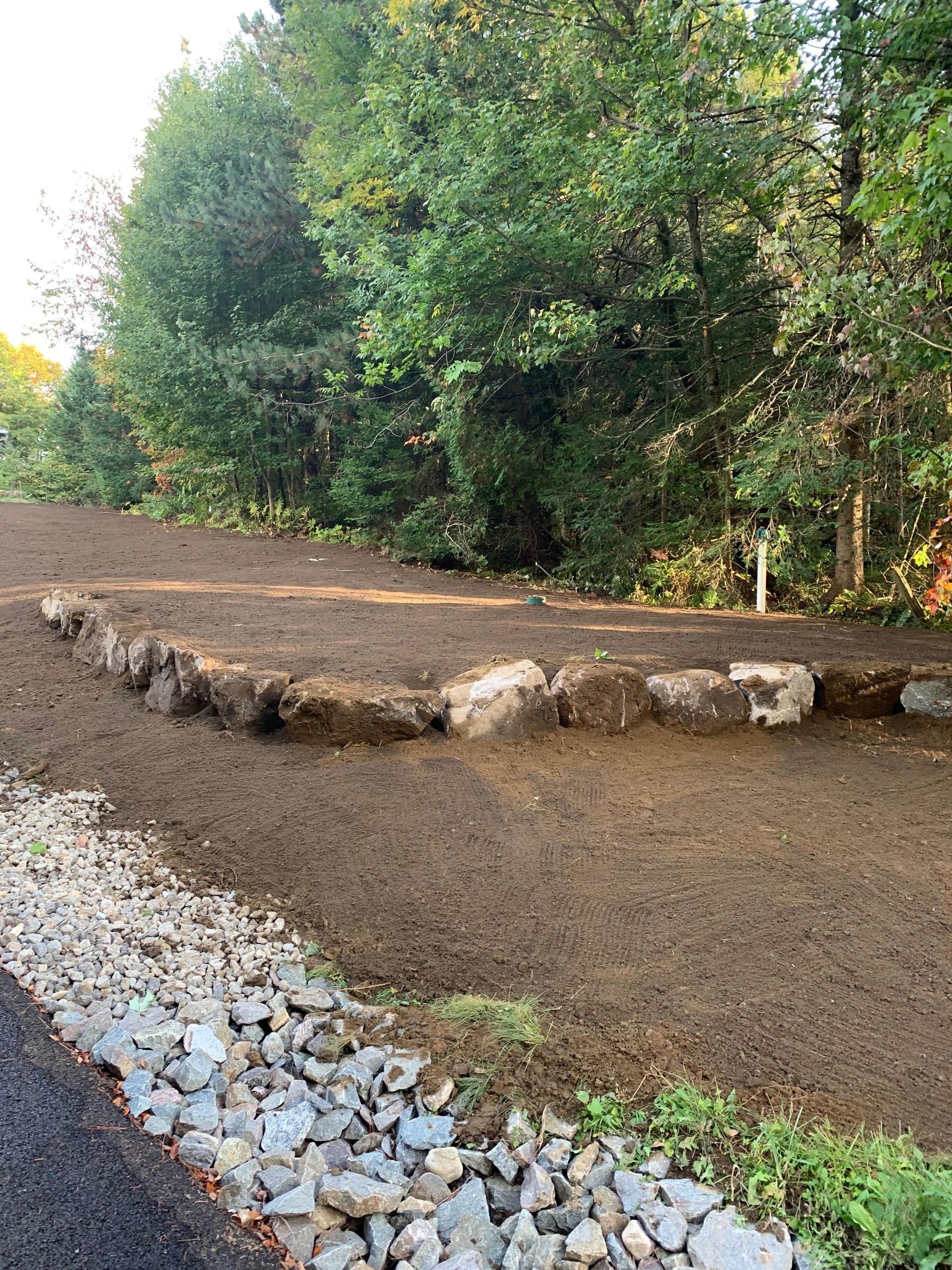 Un mur de soutènement en pierre se dresse au milieu d'une cour en terre fraîchement nivelée, bordée par un canal de drainage en gravier et un bois dense.