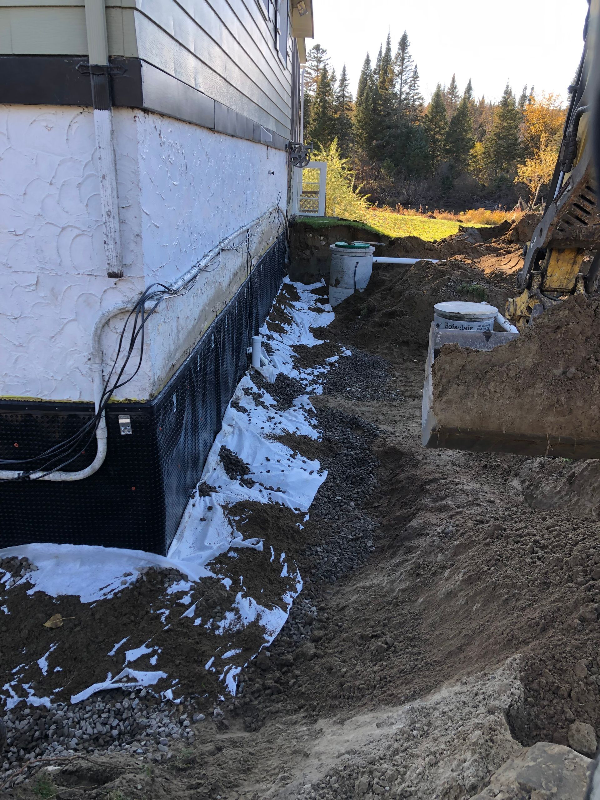 Vue latérale des fondations d'une maison en cours d'imperméabilisation par un revêtement noir et installation d'un tuyau de drainage dans une tranchée excavée.