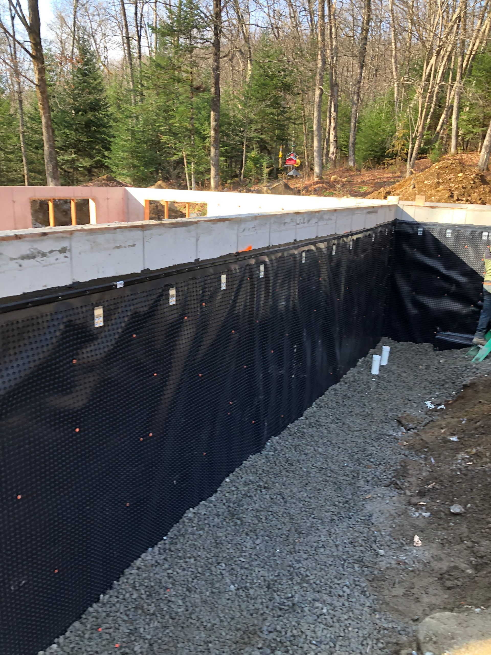 Un mur de fondation avec une membrane de drainage noire à picots installée contre du gravier et de la terre, avec des bois en arrière-plan.