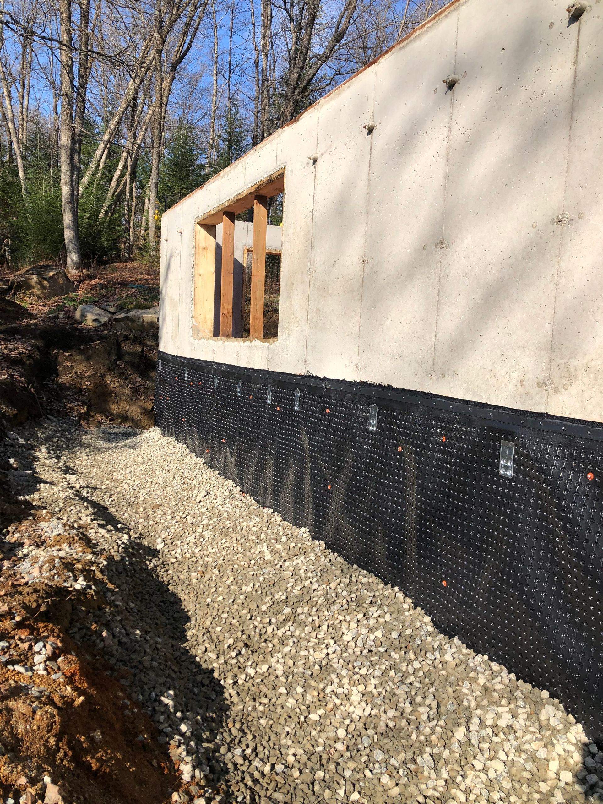 Un mur de fondation en béton avec une membrane d'étanchéité noire à picots, se détachant sur un fond de tranchée de gravier et de végétation boisée.