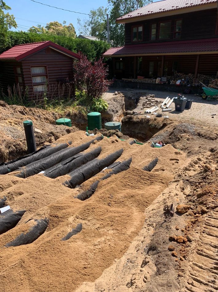 Un chantier montrant une fosse septique nouvellement installée avec des tuyaux noirs partiellement enterrés dans le sable près d'une maison.