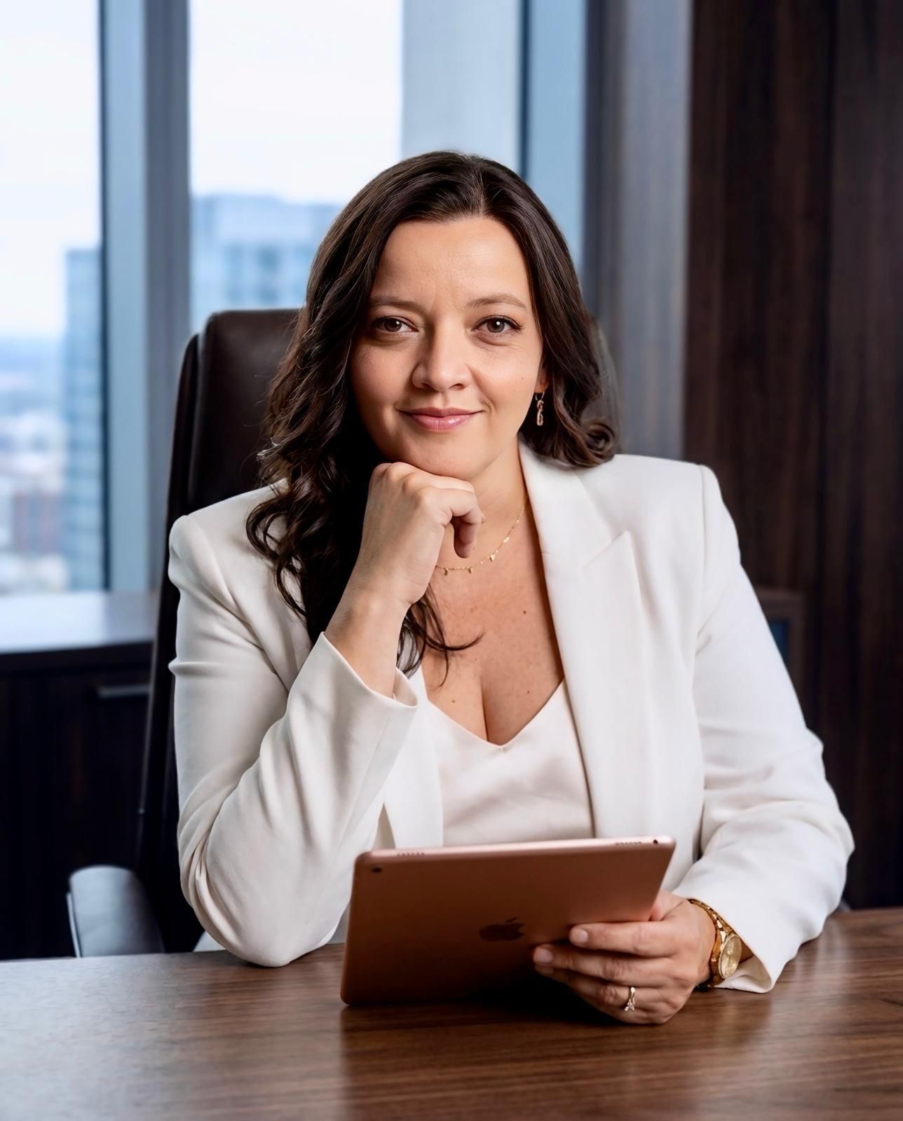 Professional woman seated at a desk in an office, holding a tablet and smiling at the camera.