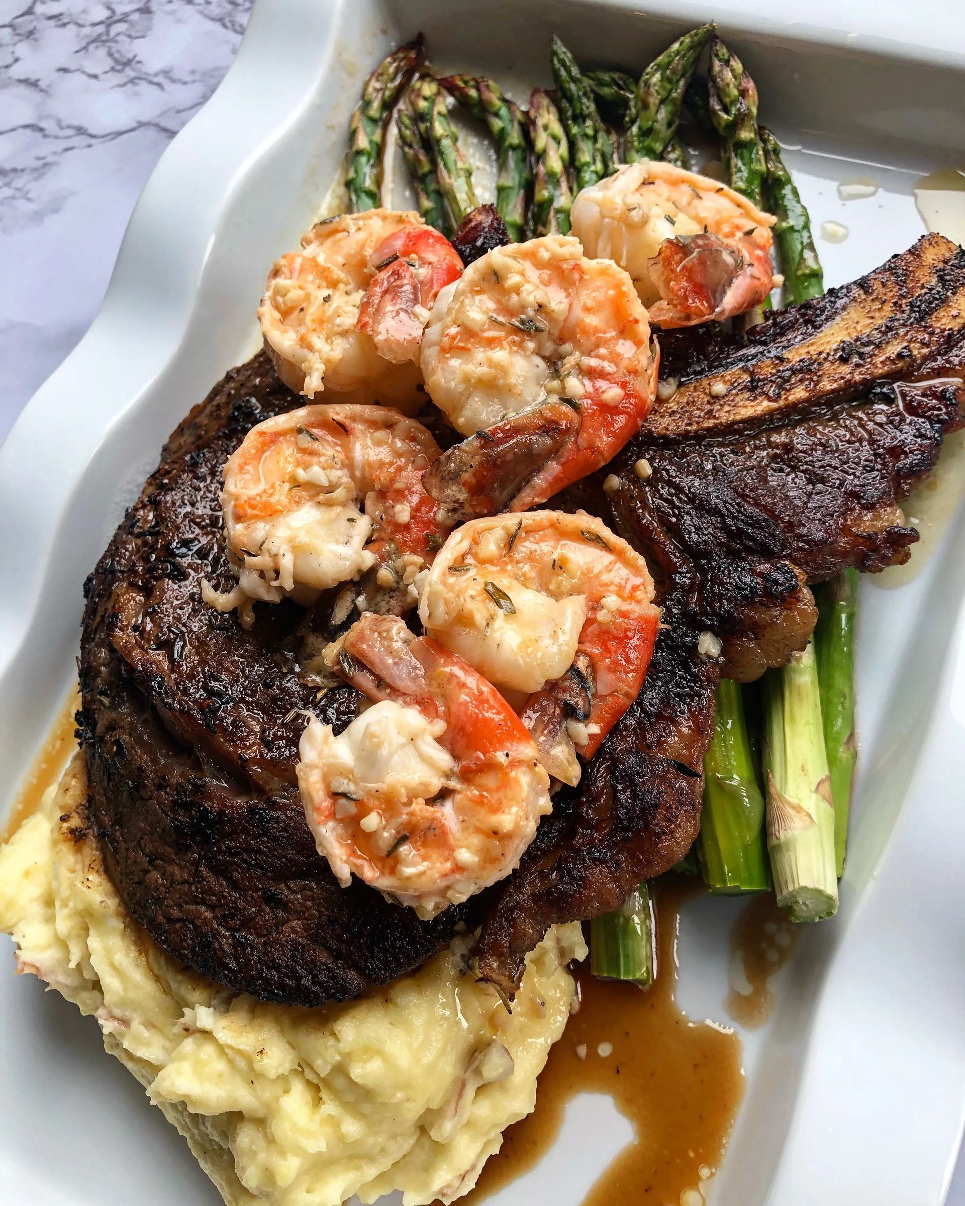 Grilled steak topped with shrimp over asparagus and mashed potatoes on a white plate