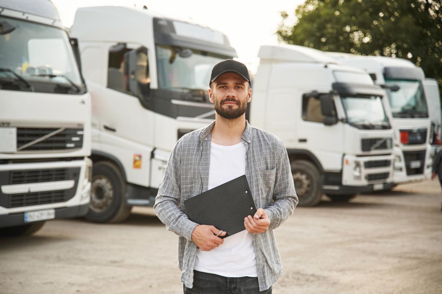 Hombre con gorra y portapapeles frente a camiones semirremolque blancos, Programa para empresa de transporte.