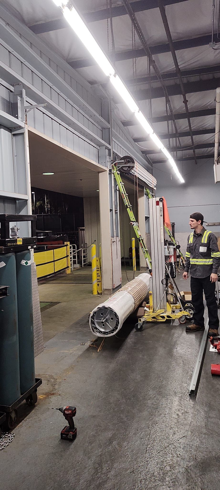 Man in safety vest observing industrial equipment, with a large cylindrical object on the floor of a warehouse.