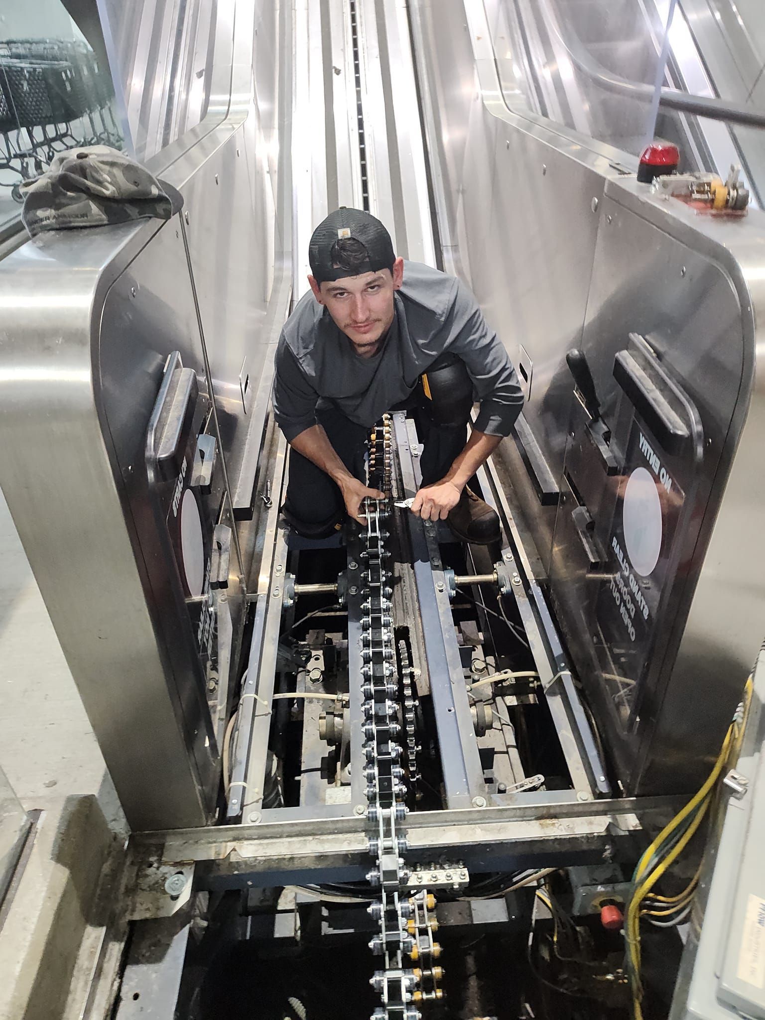 Man in gray uniform working on escalator machinery, with tools. Inside, metal structure.